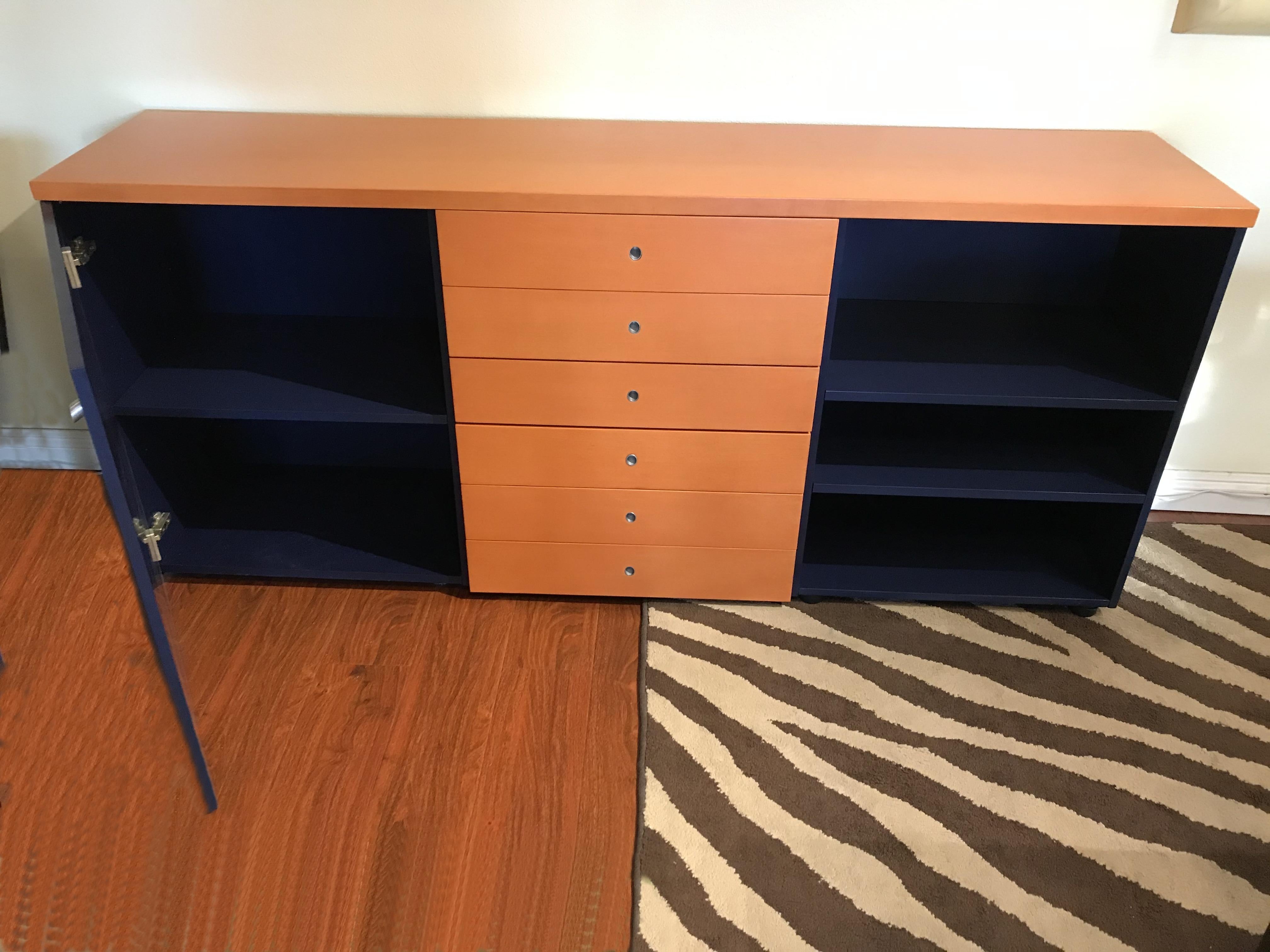 A two-toned buffet in terracotta orange and dark blue stained wood, with brushed steel accented pulls. 
It features six center drawers. The left side the door opens on 1 adjustable shelf and the right side features a little bookcase with 2