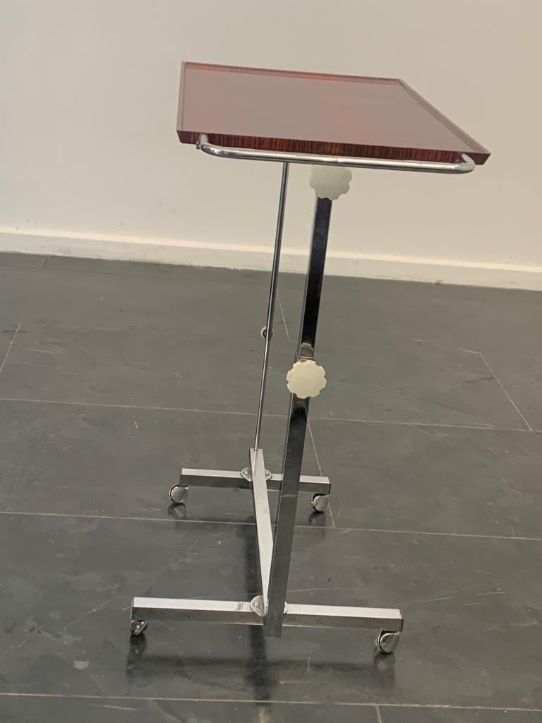 Chariot polyvalent Variett de Bremshey & Co. La table est réglable en hauteur. La table est réglable en continu d'une hauteur de 66 à 97 cm, le plateau de 40 cm x 65 peut être fermé. Métal chromé et plastique en bois de rose. Parfaitement