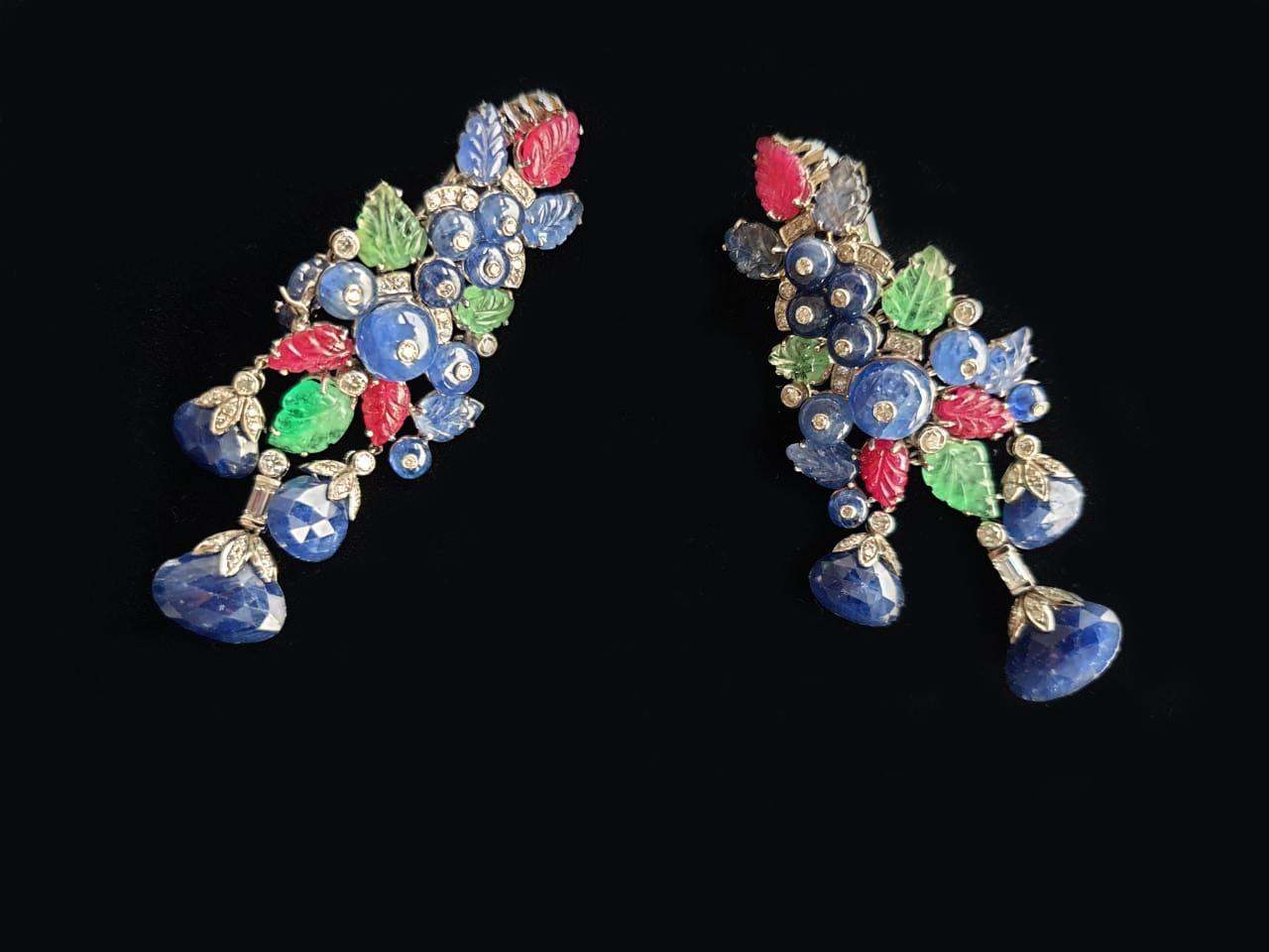 These earrings are a stunning piece of jewelry made from 18k white gold and adorned with diamonds, emeralds, rubies, and sapphires. The carved leaf designs in the gemstones add a touch of elegance and nature-inspired beauty to the overall design.