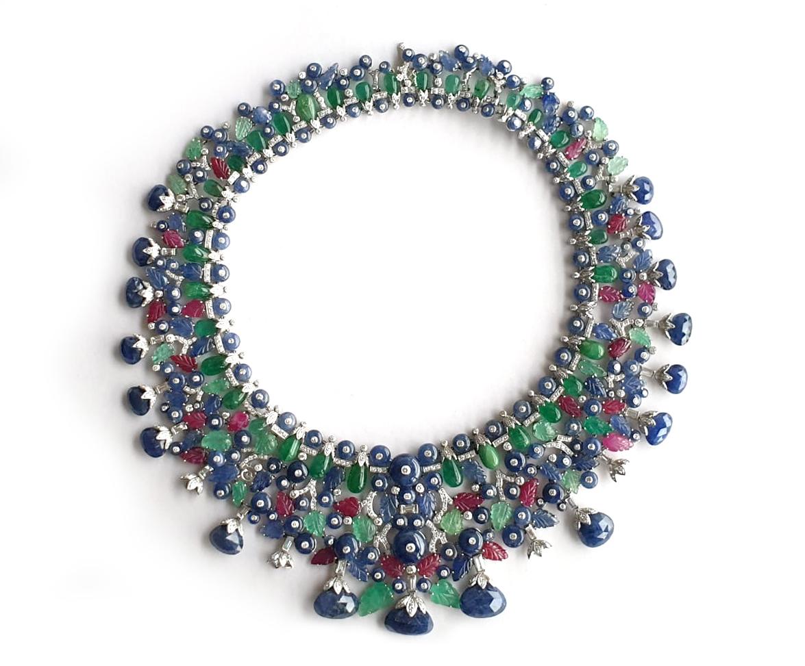 This Emerald, Ruby & Sapphire Necklace with Diamonds in 18k White Gold is a stunning piece of jewelry that features exquisite gemstones arranged in the shape of a leaf. The emerald, ruby, and sapphire are intricately carved to add texture and depth