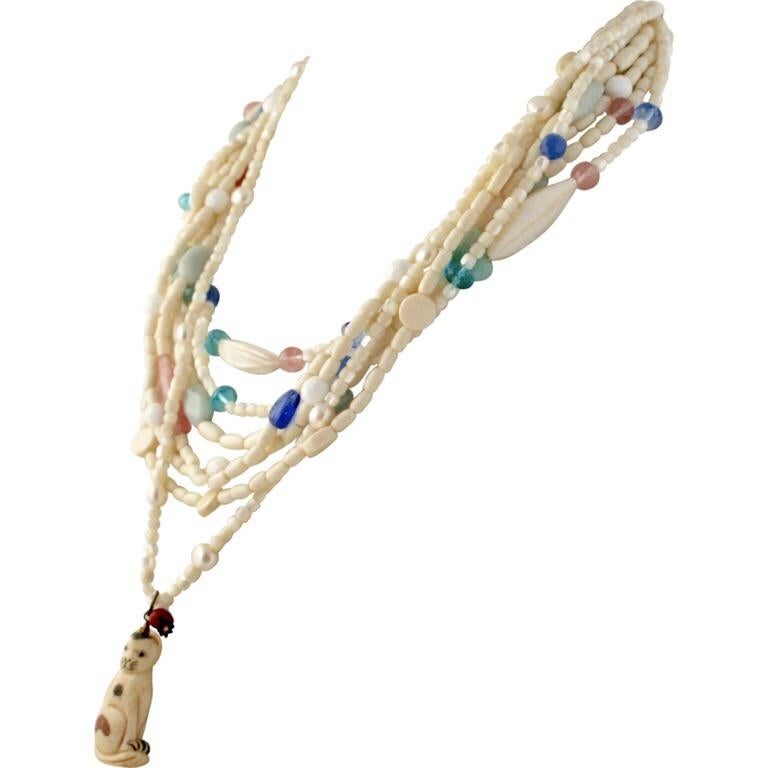 This multi-strand vintage with cat pendant necklace has pink, green, light blue stones and glass in a pastel palette along with mother-of-pearl and bone. The cat pendant is bone with color accents. The red coral bead is the cat’s toy. It is a