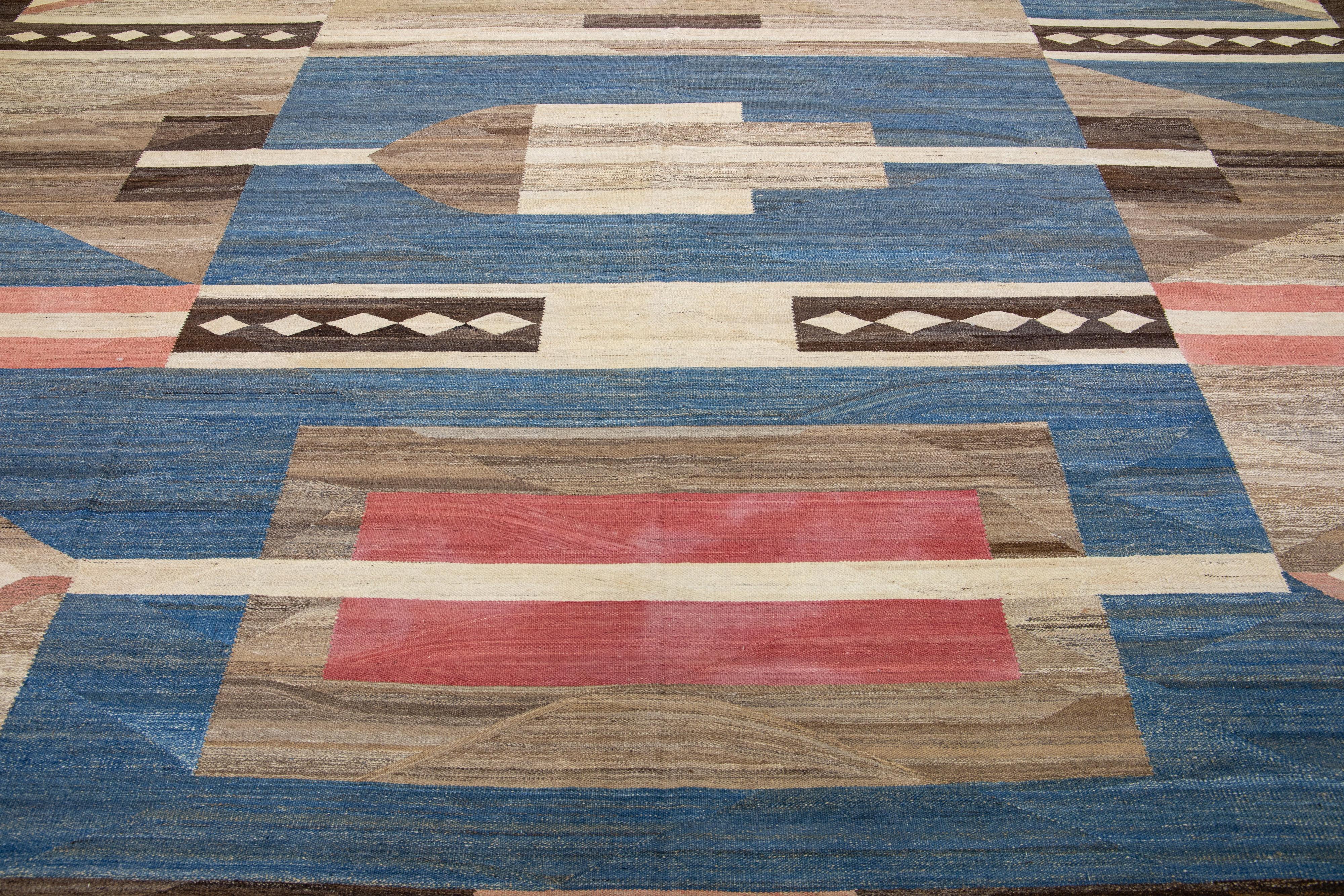 Ce tapis en laine à tissage plat présente un champ marron captivant avec des accents de couleur bleu et saumon, créant un effet géométrique contemporain et esthétiquement plaisant. Ce modèle à la mode est fabriqué à partir d'un mélange de laine
