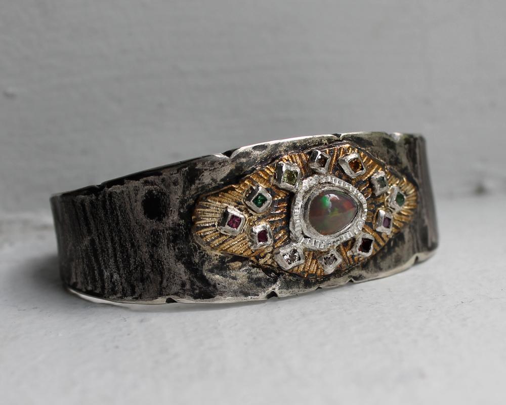 This one-of-a-kind cuff bracelet features a beautiful array of gemstones and diamonds surrounding a rare opal. The ends of the cuff have two Dreaming Muse figures. Paying homage to tradition, the cuff has been formed from scratch using heirloom