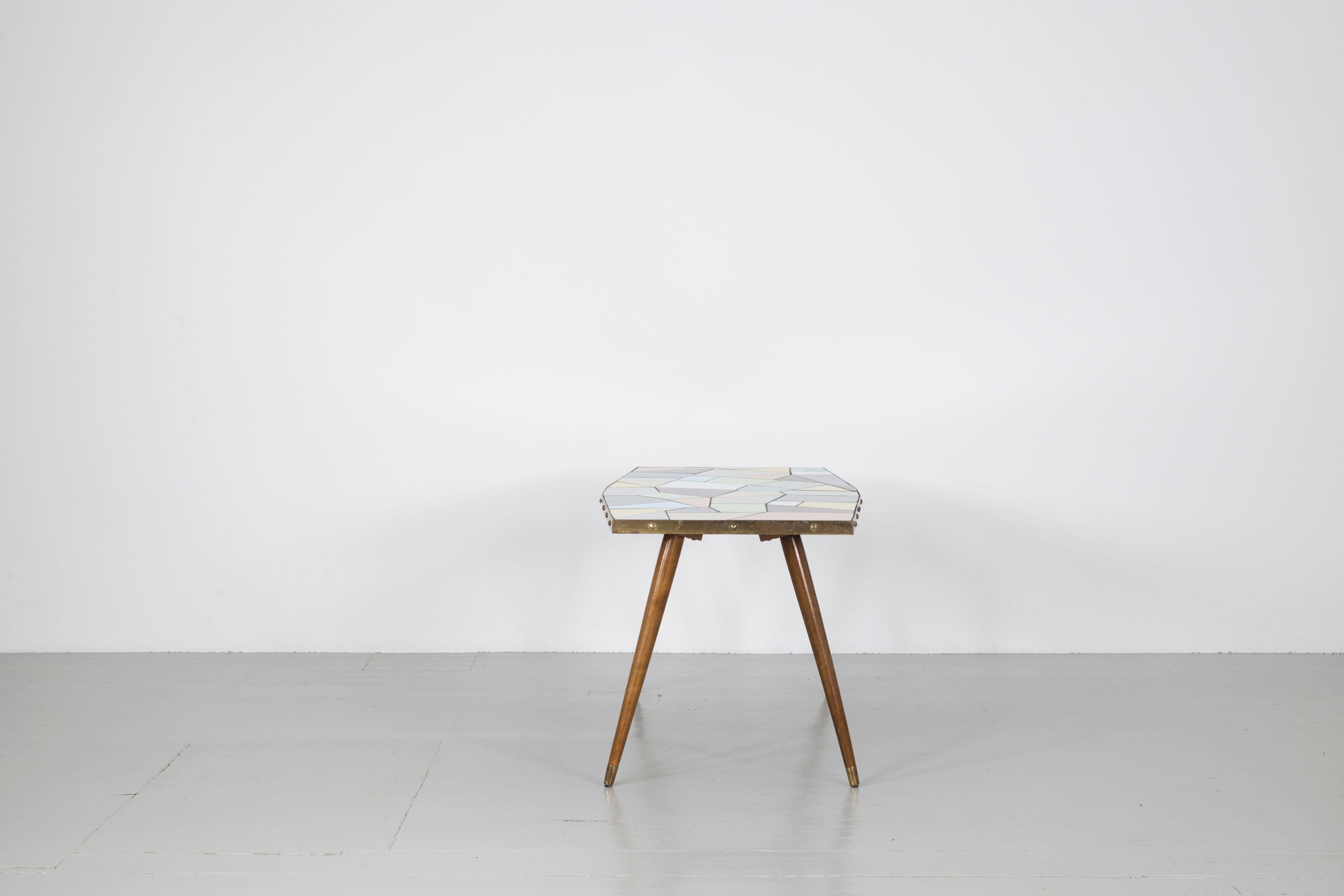 This table was made in Germany in the 1950s. The combination of robust materials and soft colour tones gives the table a special appearance. The table top is covered with tiles in soft pastel shades, white and black and surrounded by a shiny brass