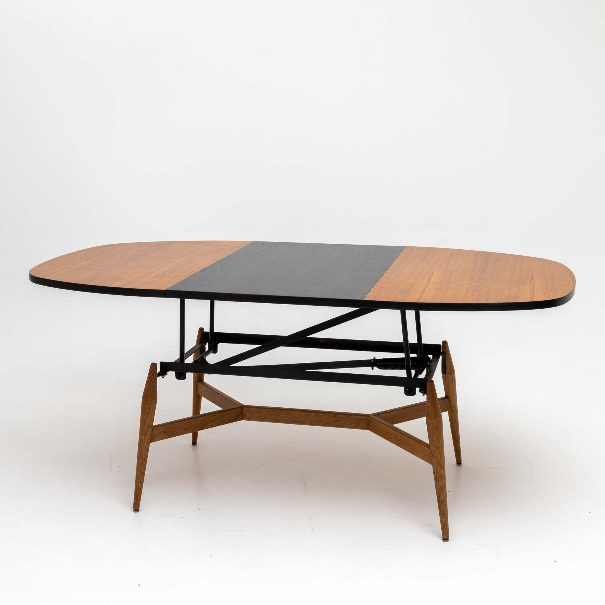 Table basse avec plateau ovale, pieds coniques et entretoise en x. En allongeant le plateau de la table et en insérant une pièce centrale (noire), elle peut être transformée en table à manger. Dimensions en extension (HxLxP) : 73,5 x 166 x 85 cm.
