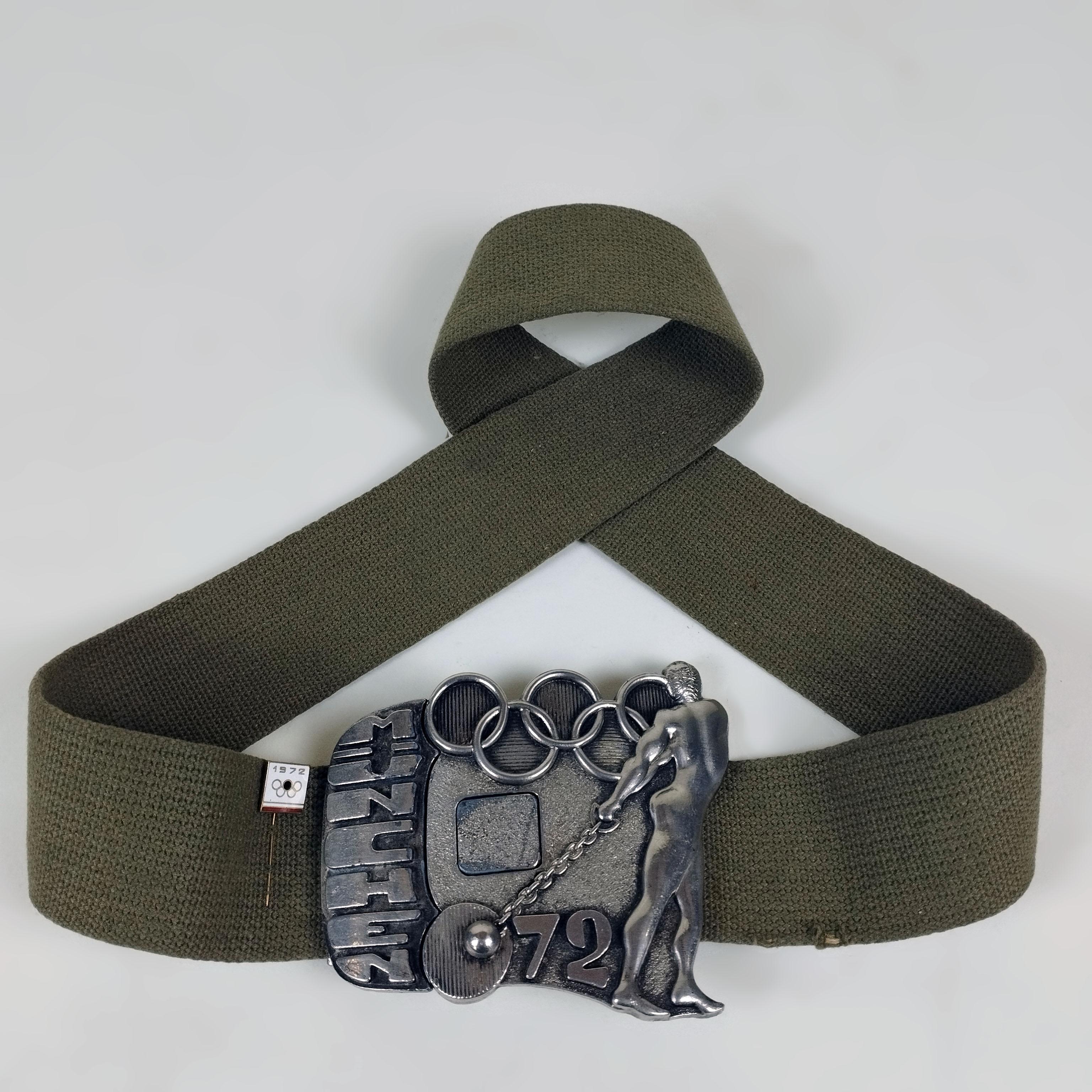 A 1972 Summer Olympics commemorative belt with metal buckle. The olive green fabric belt shows mild marks of wear. The metal buckle features the Olympic rings, the MÜNCHEN 72 words and a hammer thrower. The buckle has a metal pin attached to it.