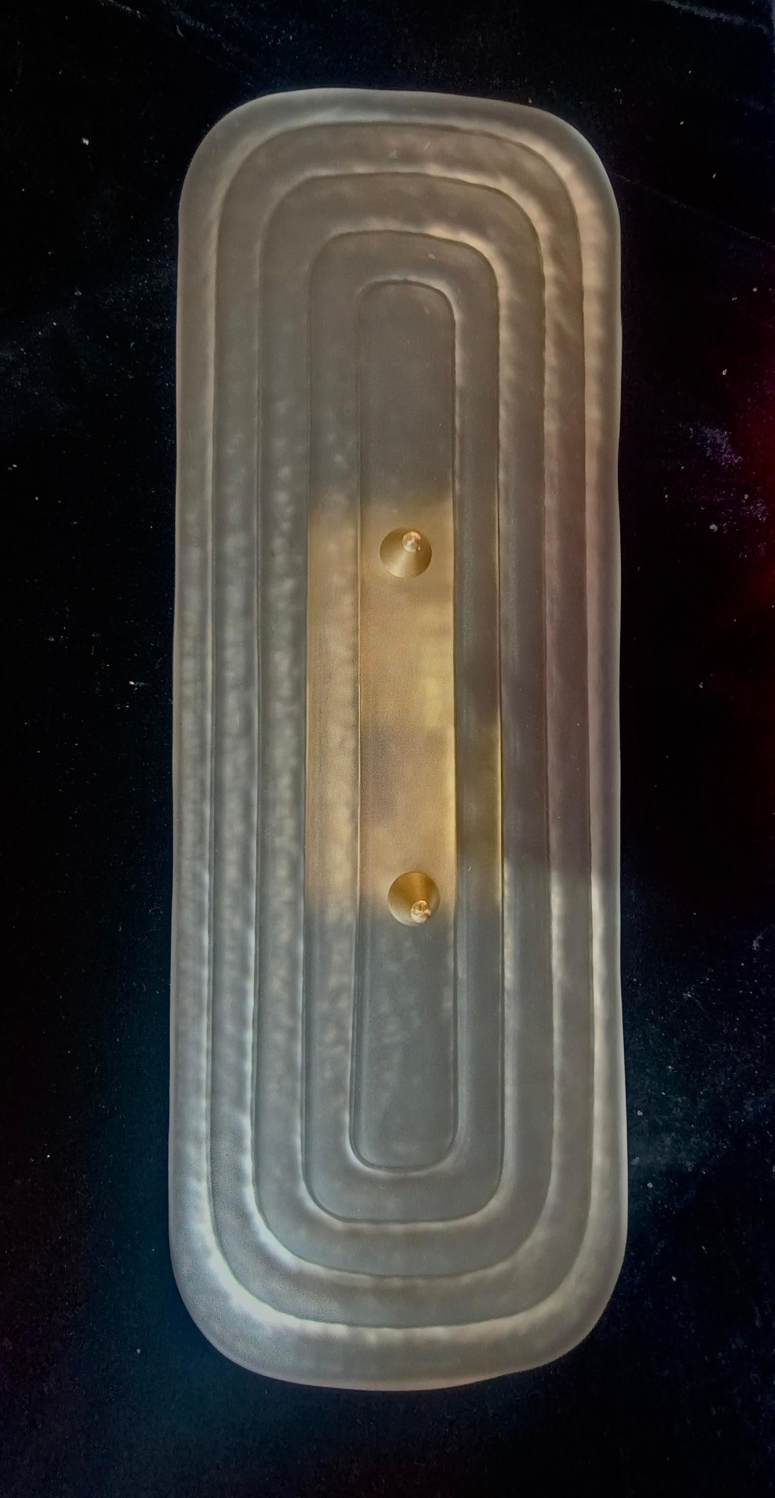 Aplique de cristal de Murano muy original con un diseño muy alargado de la placa de cristal y un color ahumado arenado único.

El aplique está compuesto por una estructura metálica de color dorado arenado a la que se ha fijado una placa de cristal