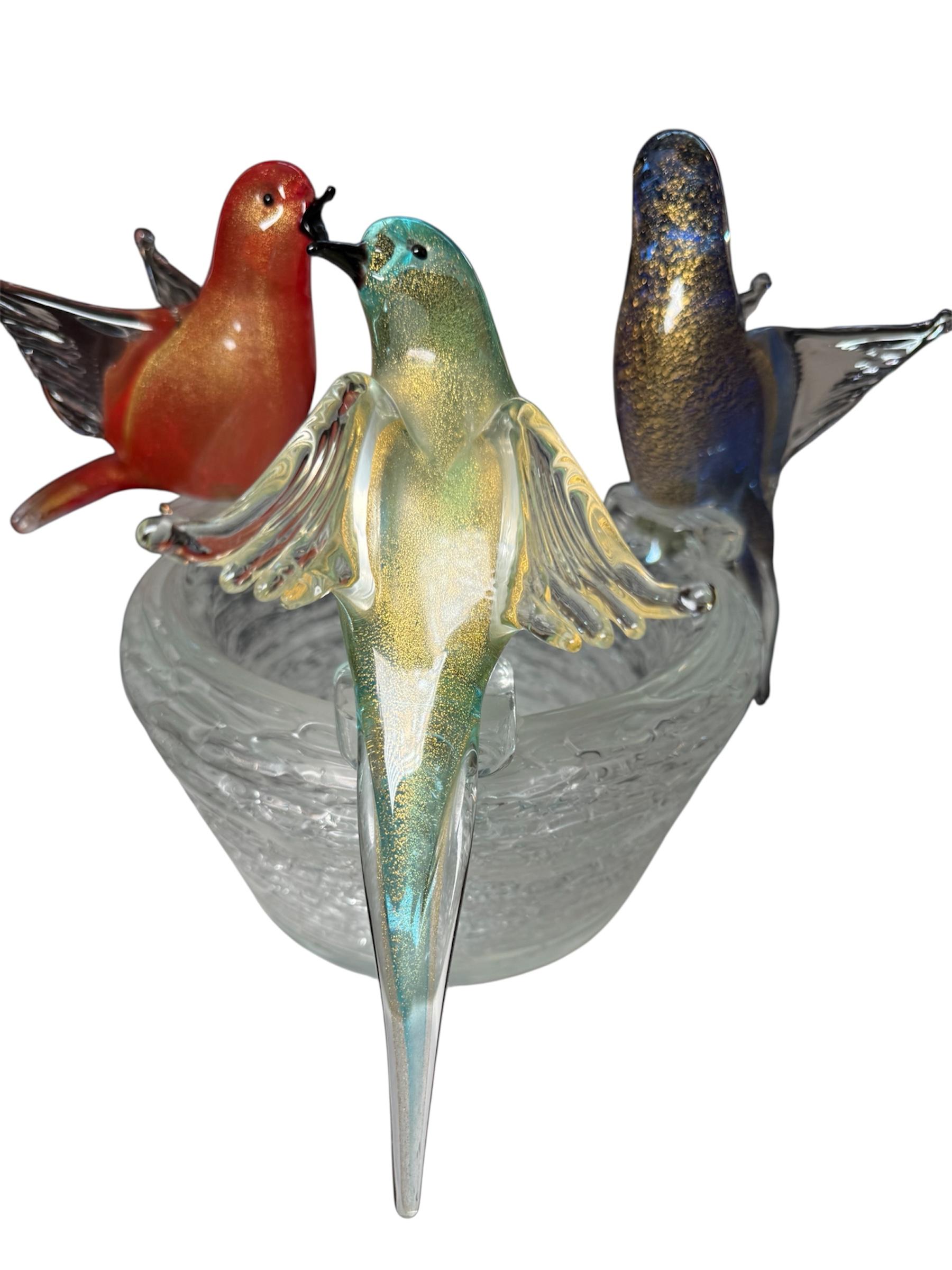 Il s'agit d'une sculpture d'art Murano représentant des oiseaux sur un nid. Il représente un nid rond en verre transparent avec trois oiseaux colorés se reposant sur sa bordure. L'oiseau orange chante et les deux autres regardent autour d'eux.