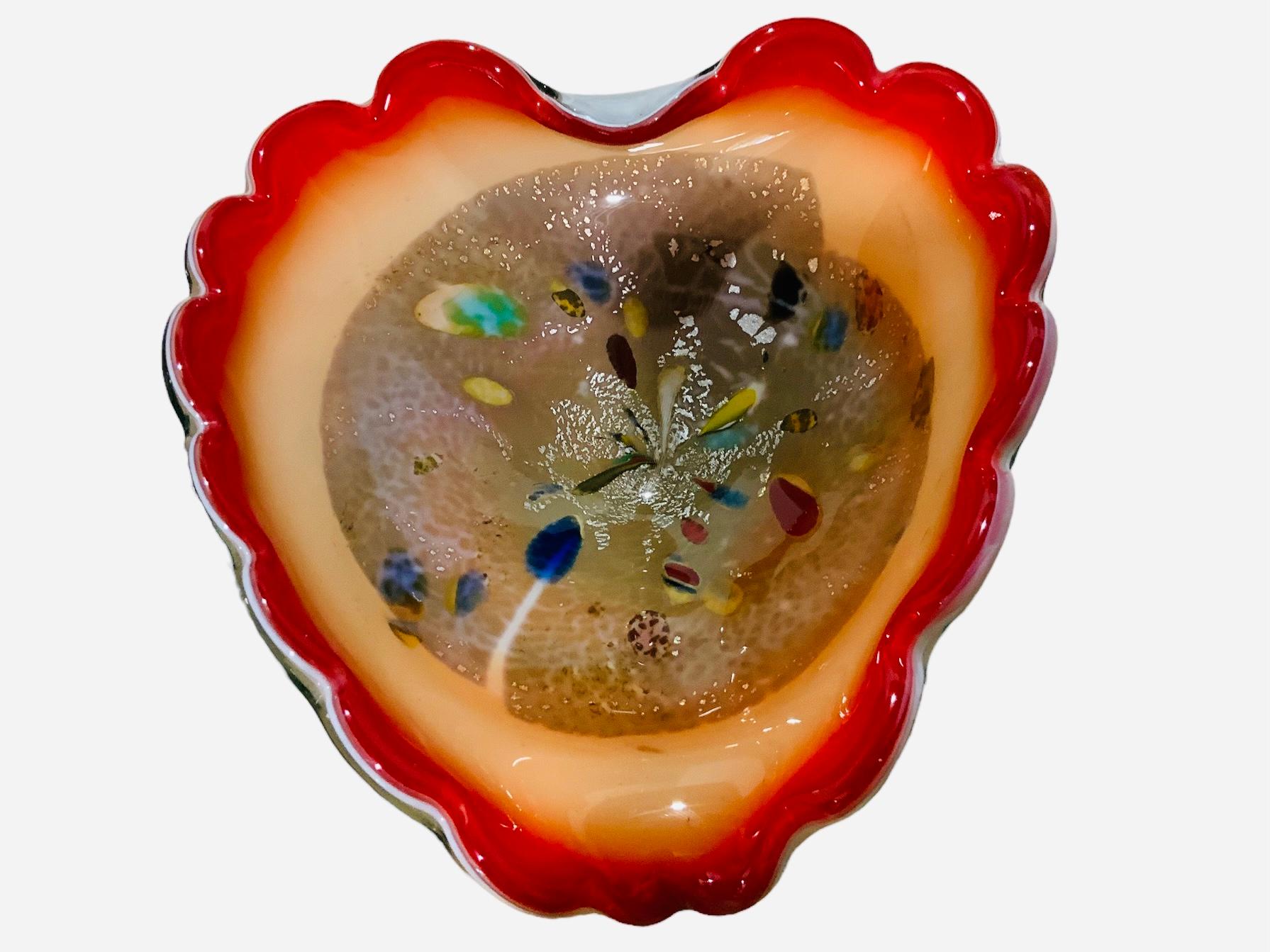 This is a Murano Art Biomorphic Glass Bowl. It depicts a heart shaped glass. It is white color in the back and in the front, its upper side is red, the middle one is light orange color and the center is clear glass enhanced with very colorful small