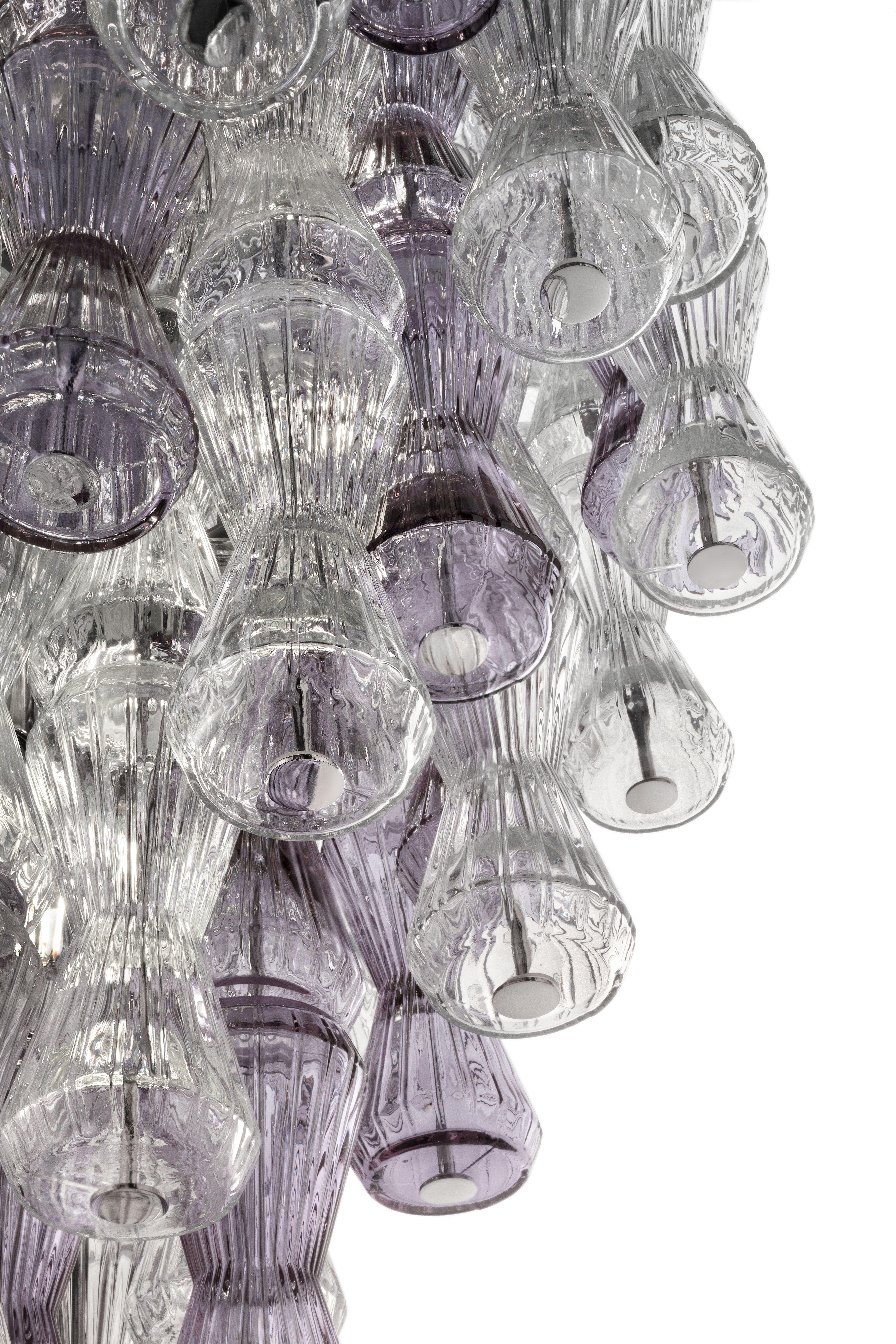 Vortici di clessidre si muovono nello spazio con la luce, una presenza danzante che attraversa ogni parte in cristallo veneziano, rivestito con l'antica tecnica Rigadin. Dalla forma sinuosa, le clessidre si ripetono e si sovrappongono, creando