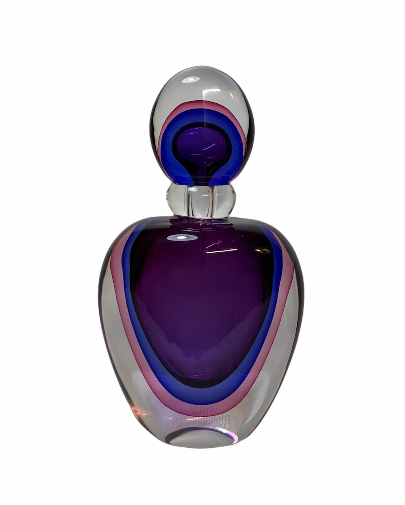 This is a Murano Crystal Perfume bottle. It is an apple shaped clear bottle that depicts U-shaped layers of purple, blue and pink colors in the center. The bottle has a wide round clear crystal neck. Its oval stopper follow the same pattern of