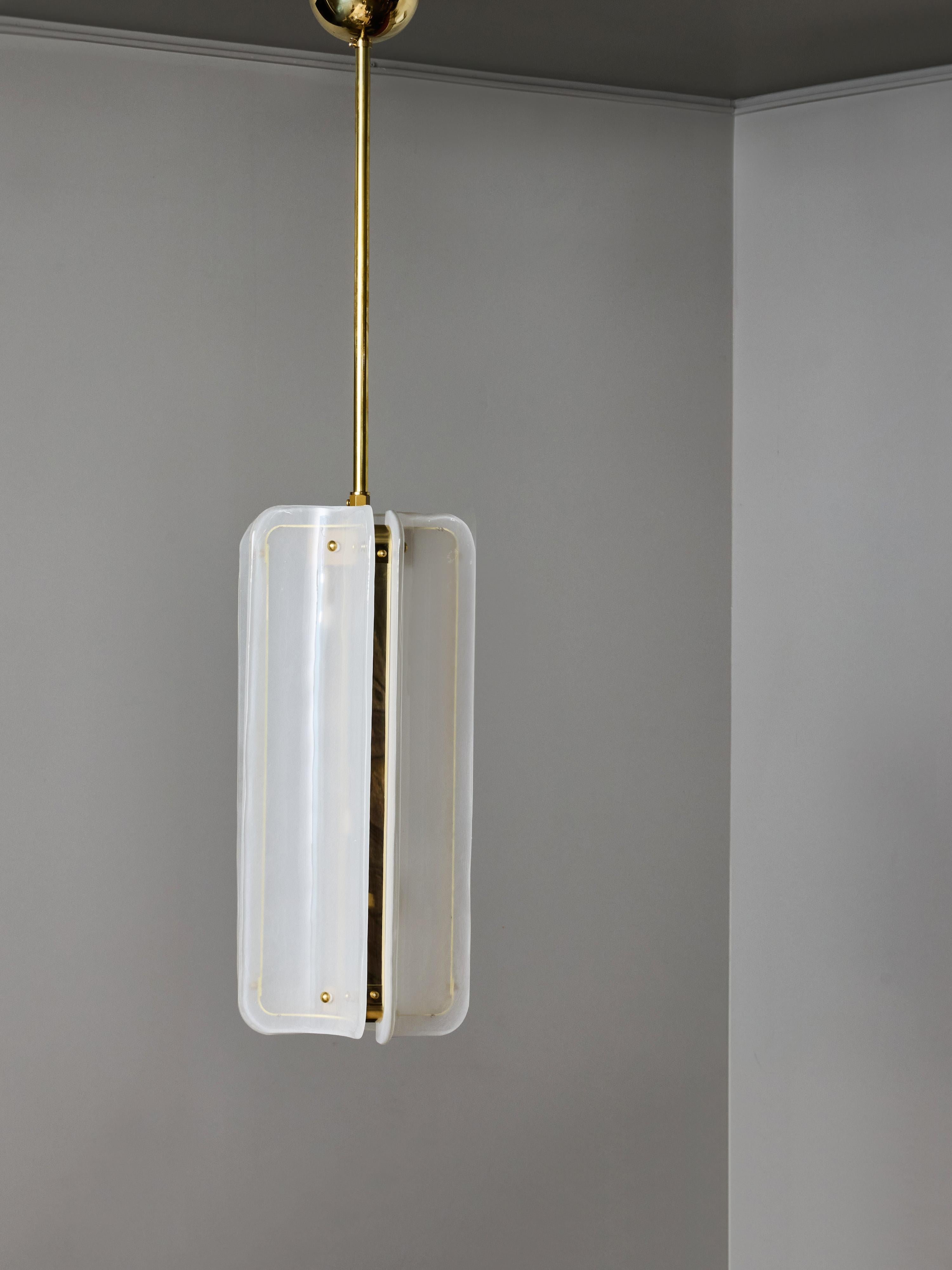 Una sofisticata lampada a sospensione moderna, caratterizzata da un assemblaggio scultoreo di pannelli di vetro liscio montati intorno a una struttura centrale in ottone. La lampada è composta da quattro elementi in vetro curvati verticalmente,