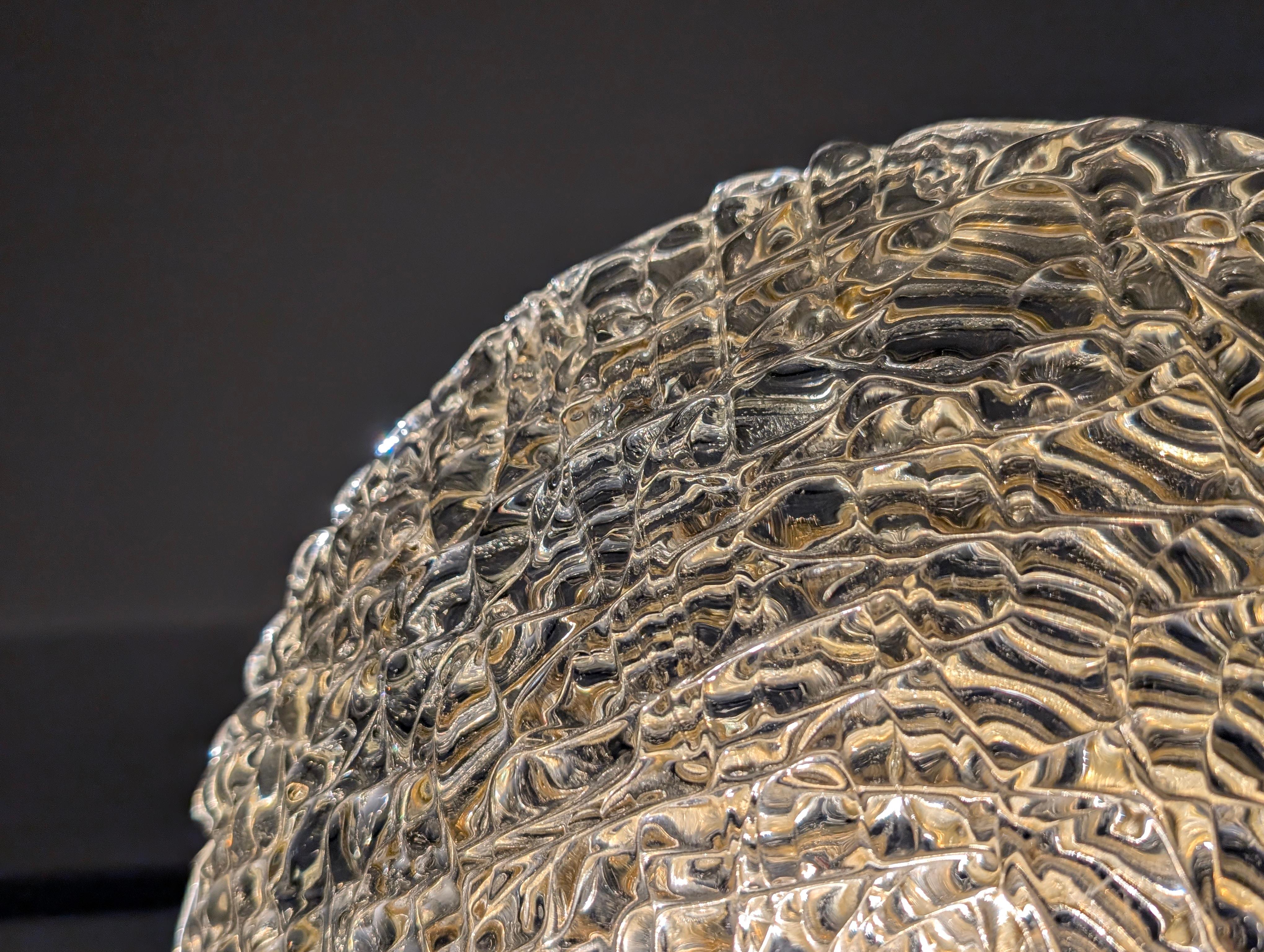 Aplique de pared circular de cristal de Murano grueso y texturado, sostenido por un estilizado soporte tubular de latón, fabricado en la década de 1970 con una característica superficie rugosa. Este cristal está directamente relacionado con la