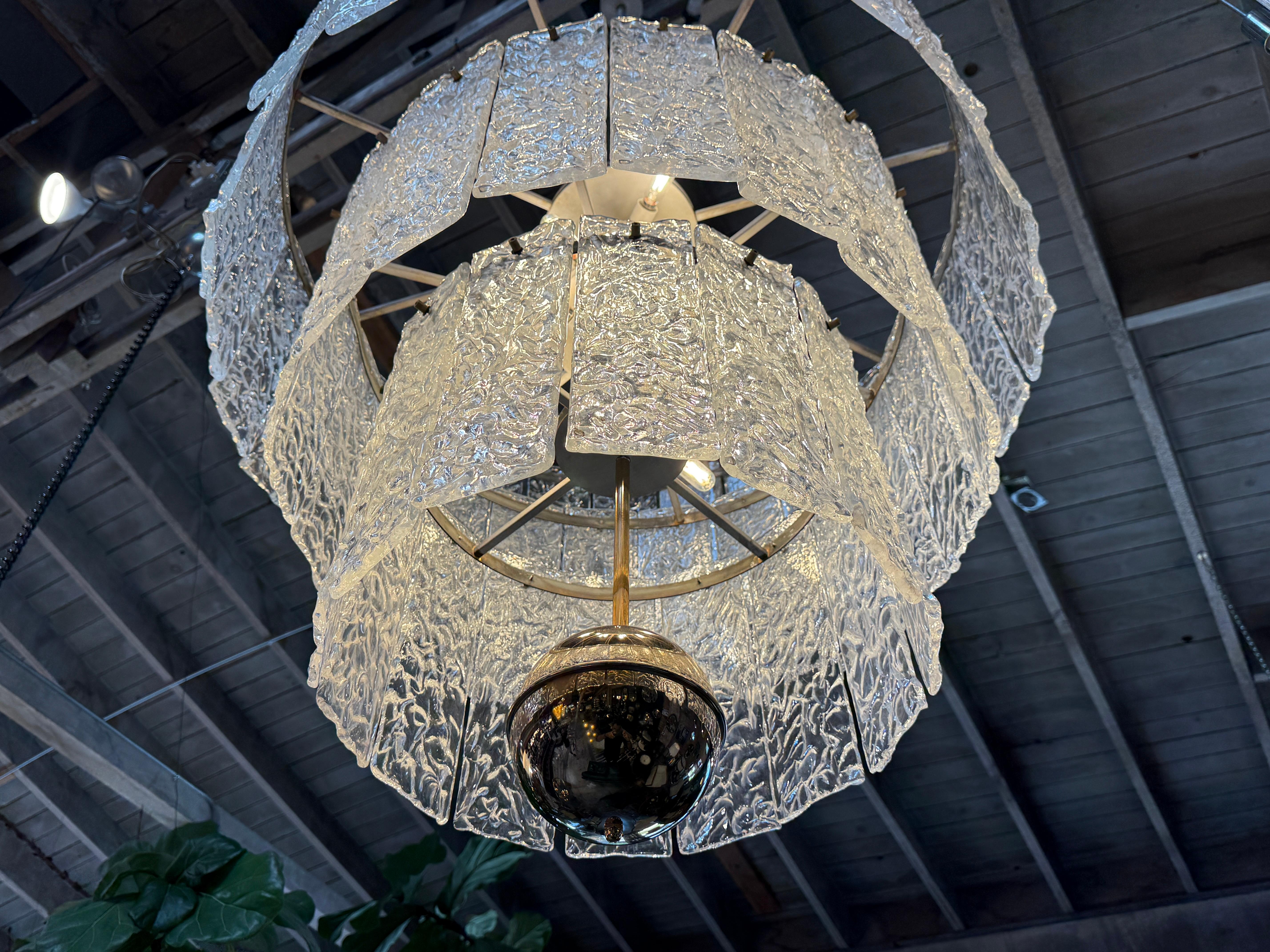 This exquisite chandelier is a mid-century modern Murano glass fixture, known for its distinctive tiered design and elegant craftsmanship. The chandelier is composed of multiple rectangular, textured glass panels that cascade down in concentric