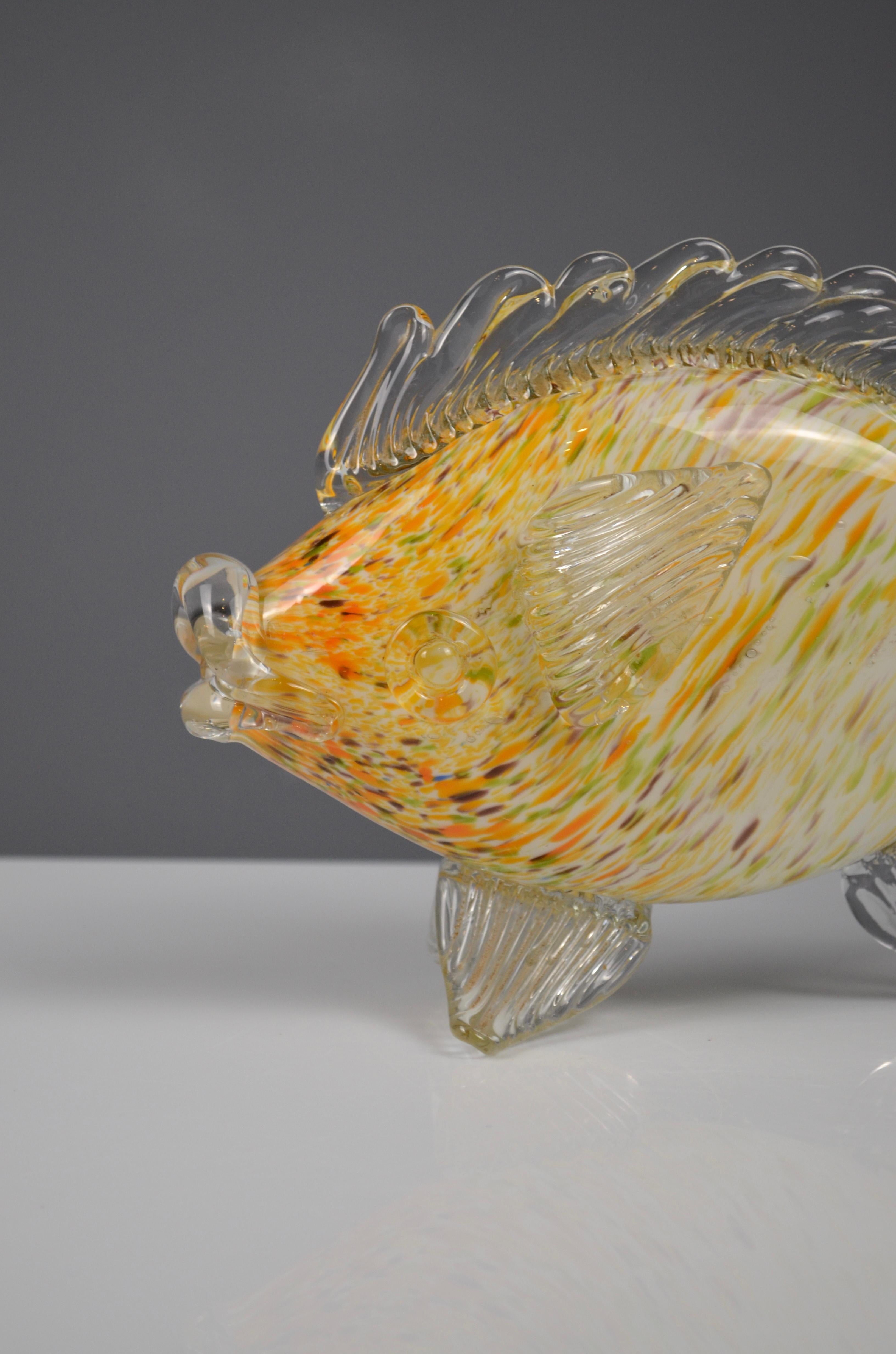 Elegant handcrafted Murano glass fish sculpture.
This decorative piece features a vibrant body with yellow, orange, and white speckles, complemented by clear glass fins and tail. The organic form and fluid transparency highlight the exceptional
