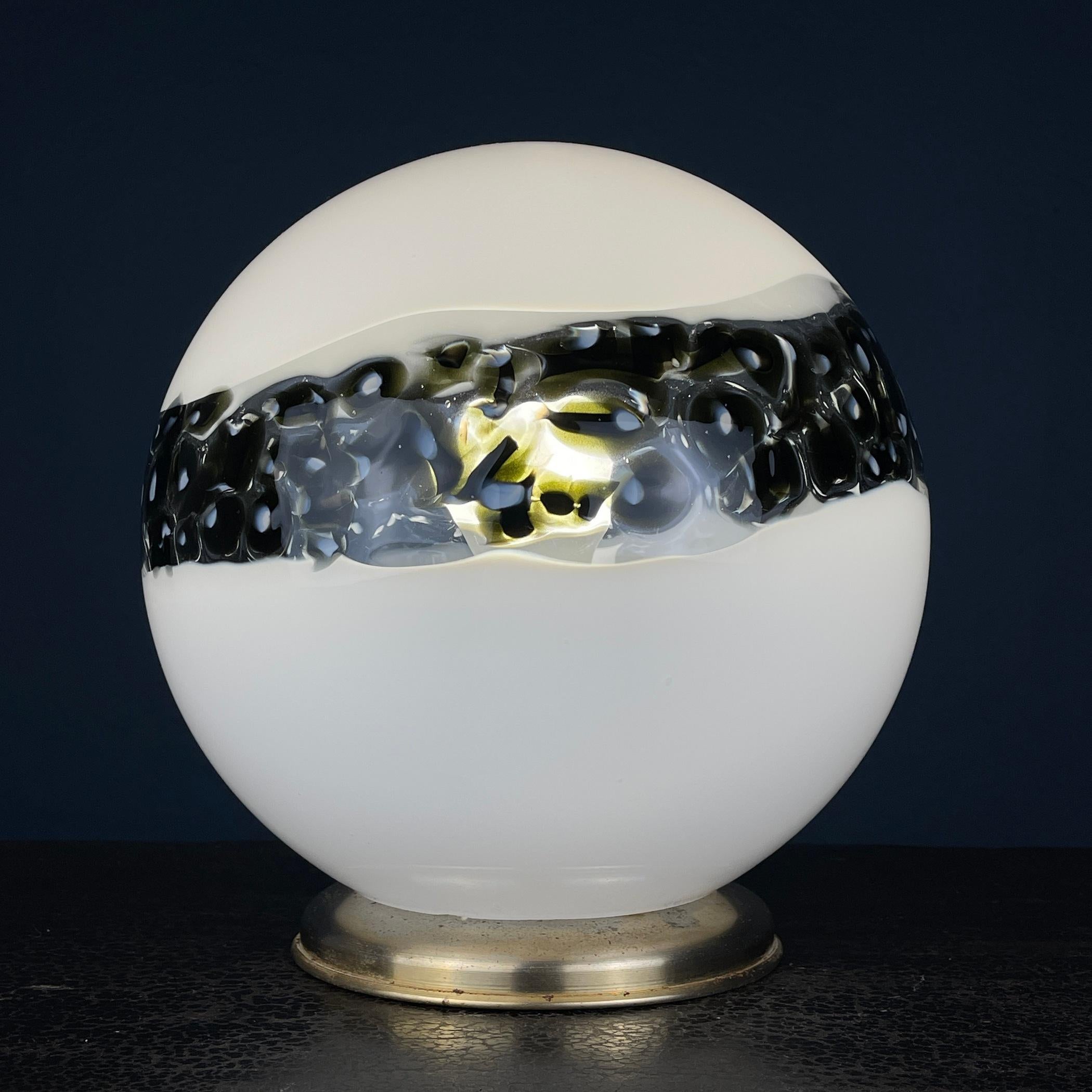 This is a splendid Murano glass table lamp from Italy, crafted in the 1970s during the Mid-century Modern era. The lamp boasts a unique and elegant design, featuring a spherical white Murano glass shade accented with a striking black stripe.

The