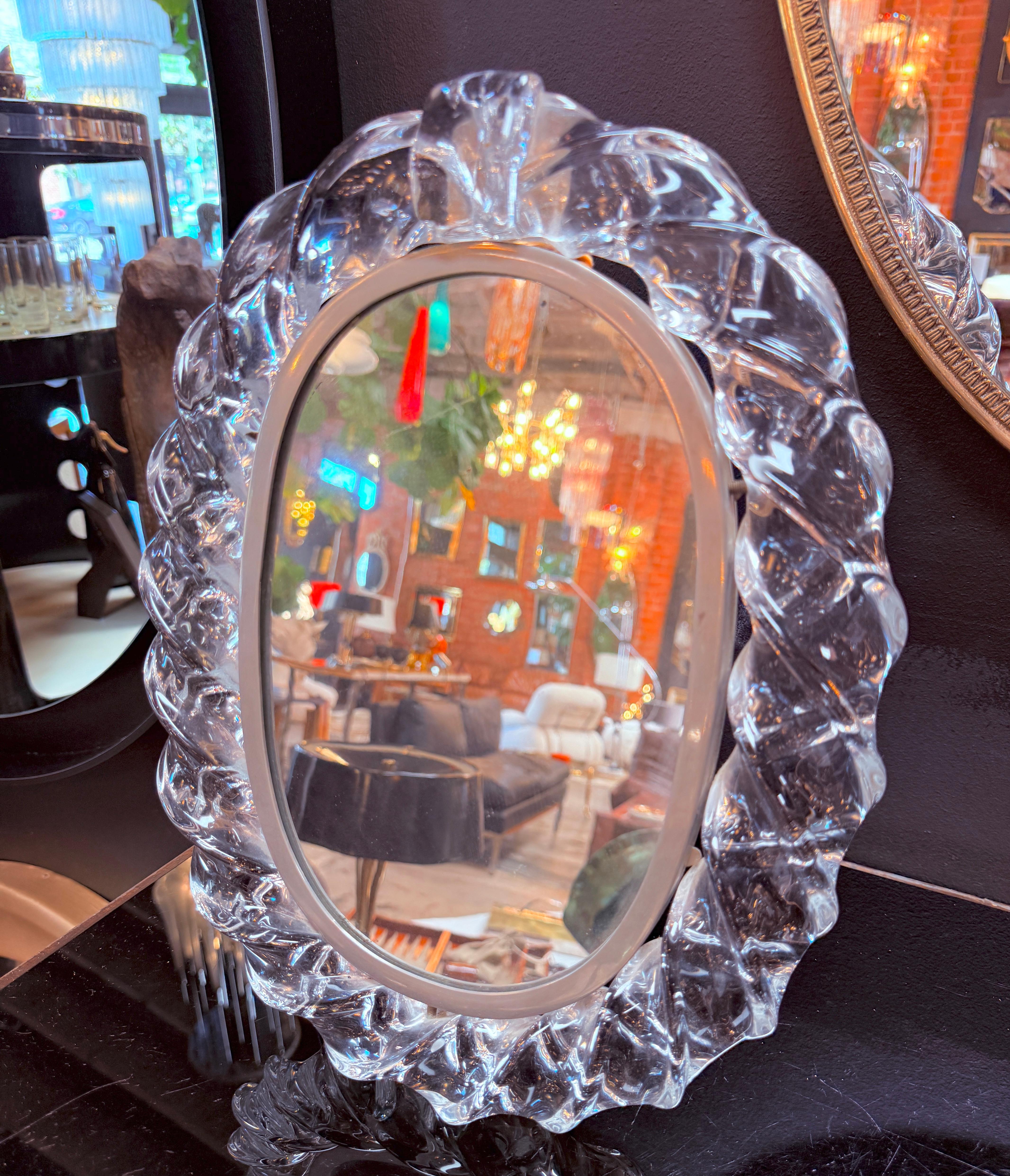 This exquisite Murano table mirror is a statement piece of Italian craftsmanship. Featuring a beautifully hand-blown crystal glass frame with intricate leaf-like detailing, it reflects the timeless artistry of Murano glassmakers. The oval mirror is