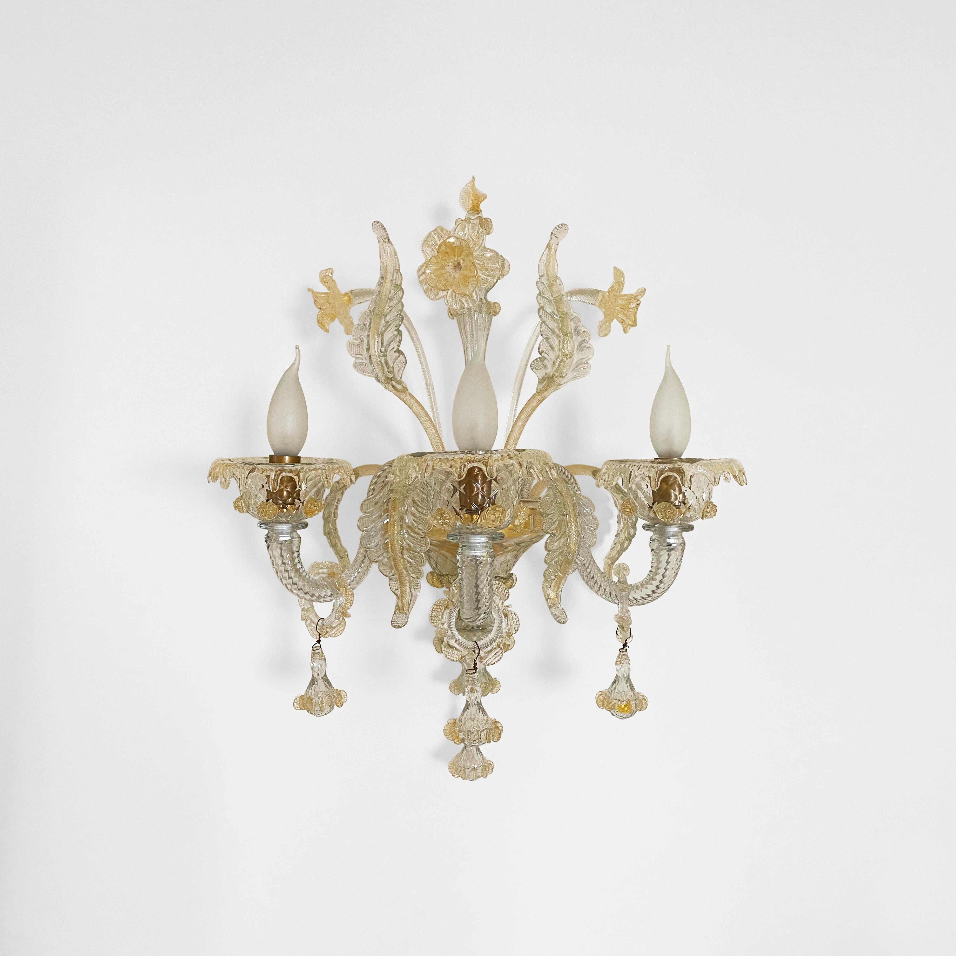 Un élégant lustre mural en verre vénitien à trois lumières de couleur cristal avec des décorations en feuilles d'or 24K. 

Les feuilles et les fleurs en verre jaillissent du sommet comme un bouquet, tandis que les trois bras tiennent les ampoules du