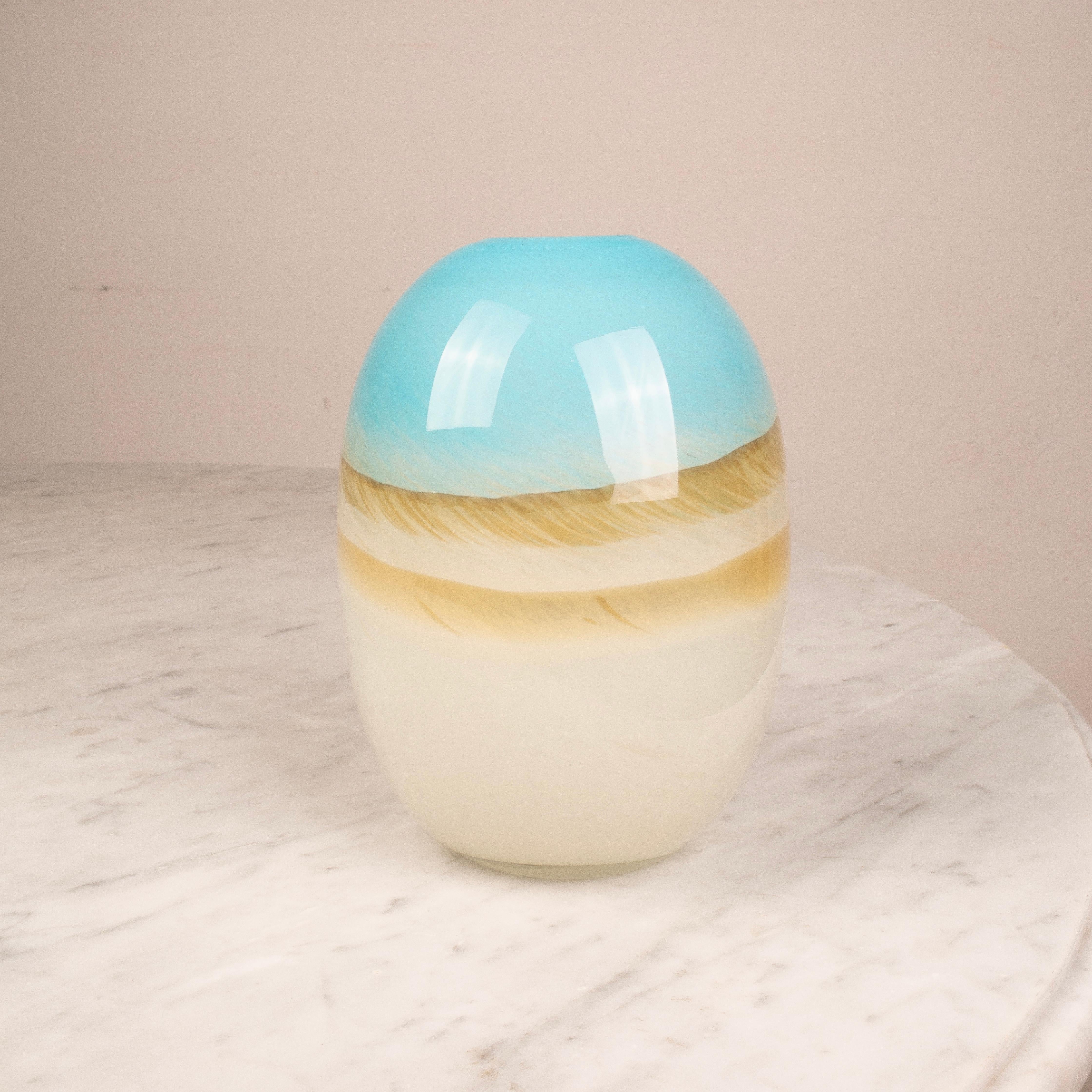 Hand-blown glass vase featuring a smooth, rounded silhouette and a refined gradient of colors. The top portion displays a vibrant sky blue hue, fading gently into warm sandy tones and finally into a soft ivory base. The glossy finish enhances the