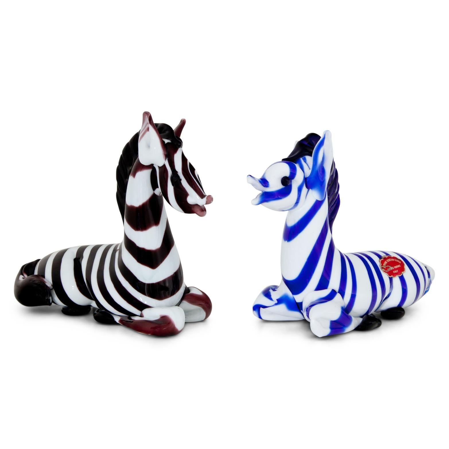 Two zebras out of Murano glass, one is red and white striped, the other one blue and white. Both are labeled “Genuine Venetian Glass, Made in Murano, Italy”.
