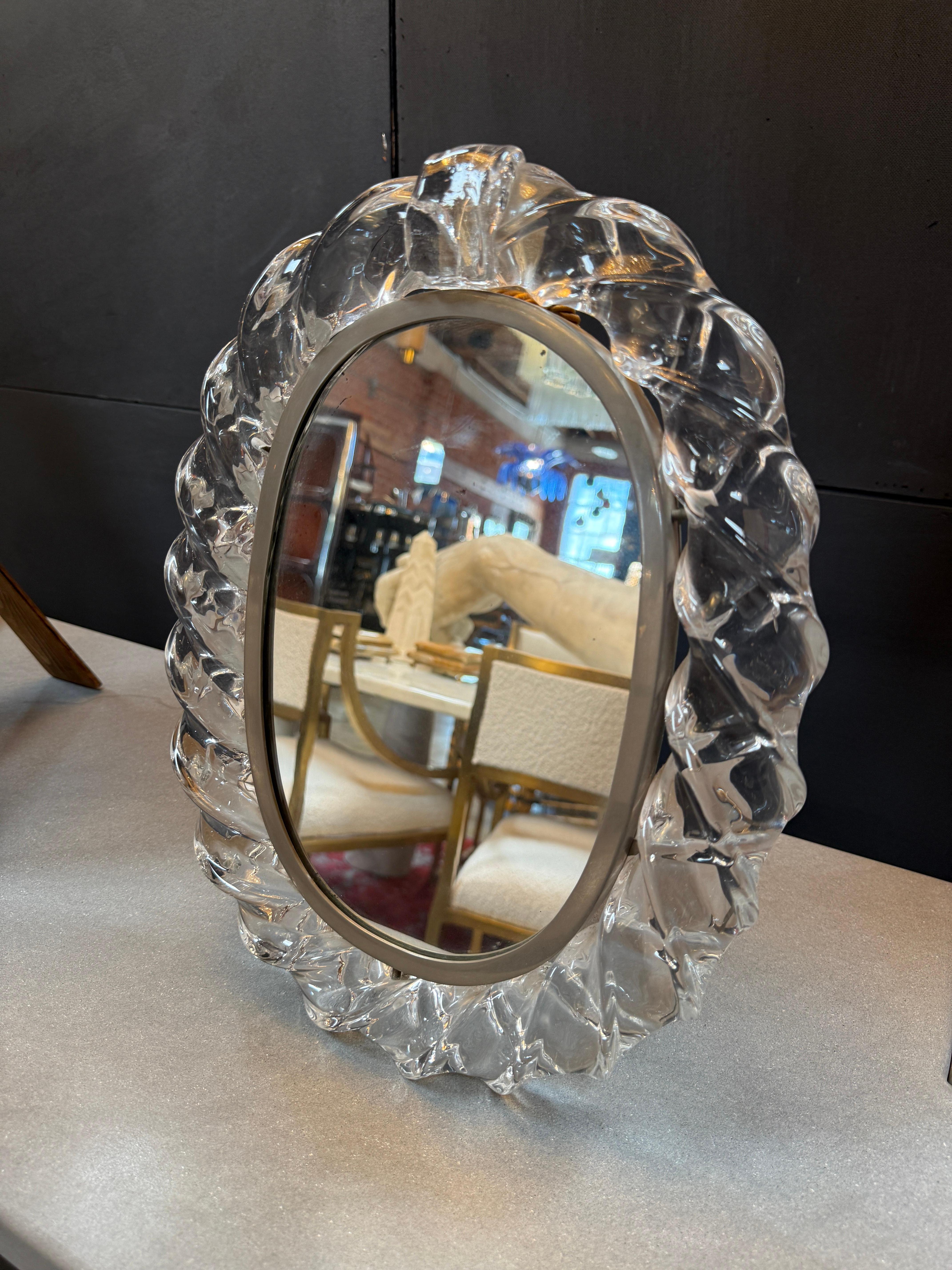This is a table mirror with a sculptural glass frame, blending elegance with artisanal craftsmanship.

The frame is made from clear, hand-cast glass, featuring a twisted, ribbon-like design that gives it a sense of movement and depth.

The glass is