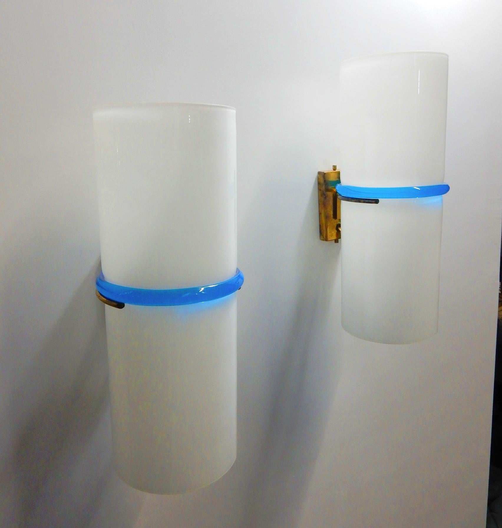 Pair of 1950s art glass lamp sconces.
Designed by Vistosi, Murano Italy.
Cased white glass cylinder belted with a gorgeous sapphire blue glass band.
Shades held firmly in place via a minimalist brass wall bracket. 
Each takes a standard light