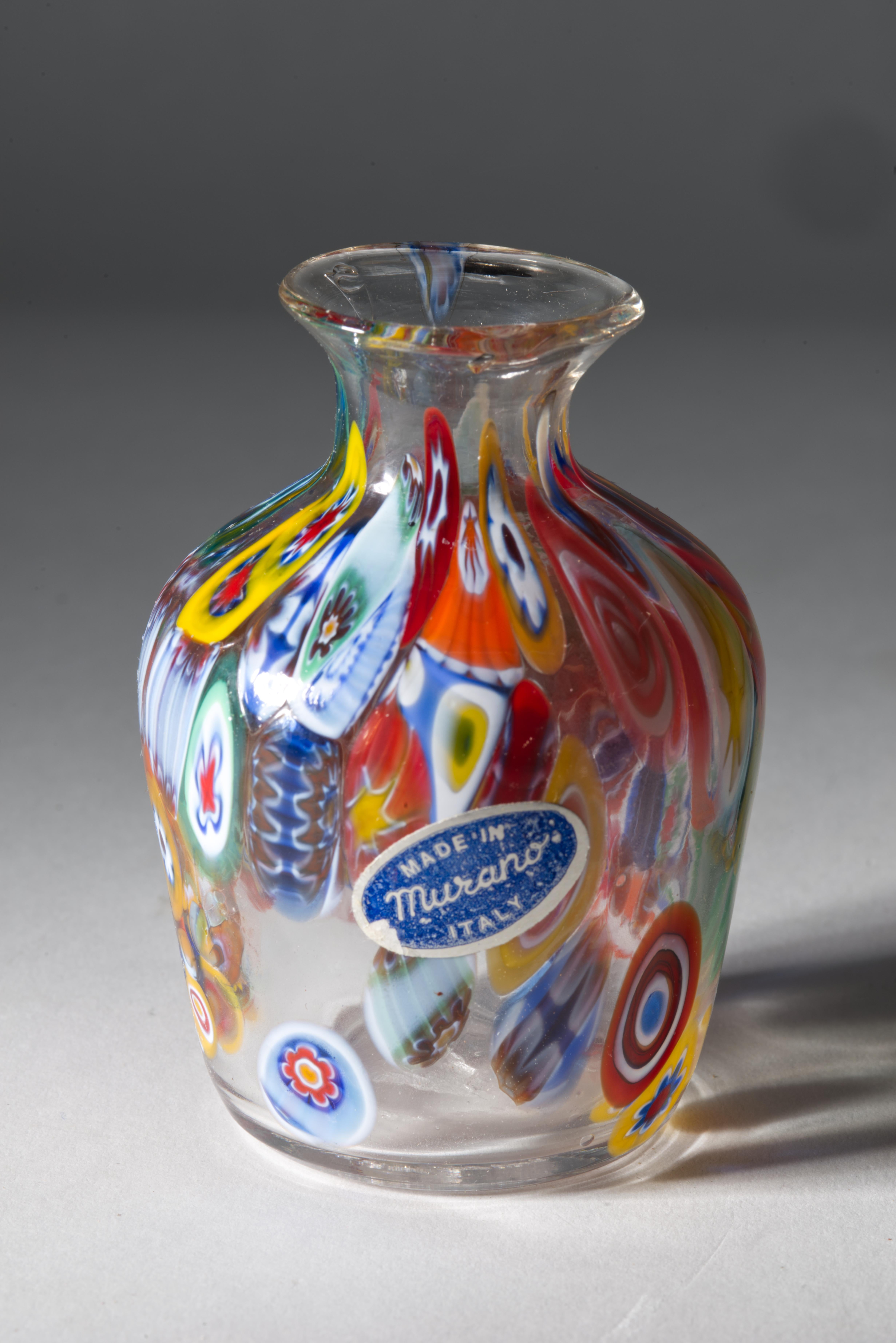  Eine ungewöhnliche Knospenvase aus Murano-Glas mit bunten Millefiori, die entlang der Linien der Vase auf klarem Glas fließen. 

Die Vase ist mit einem ovalen blauen Folienetikett 