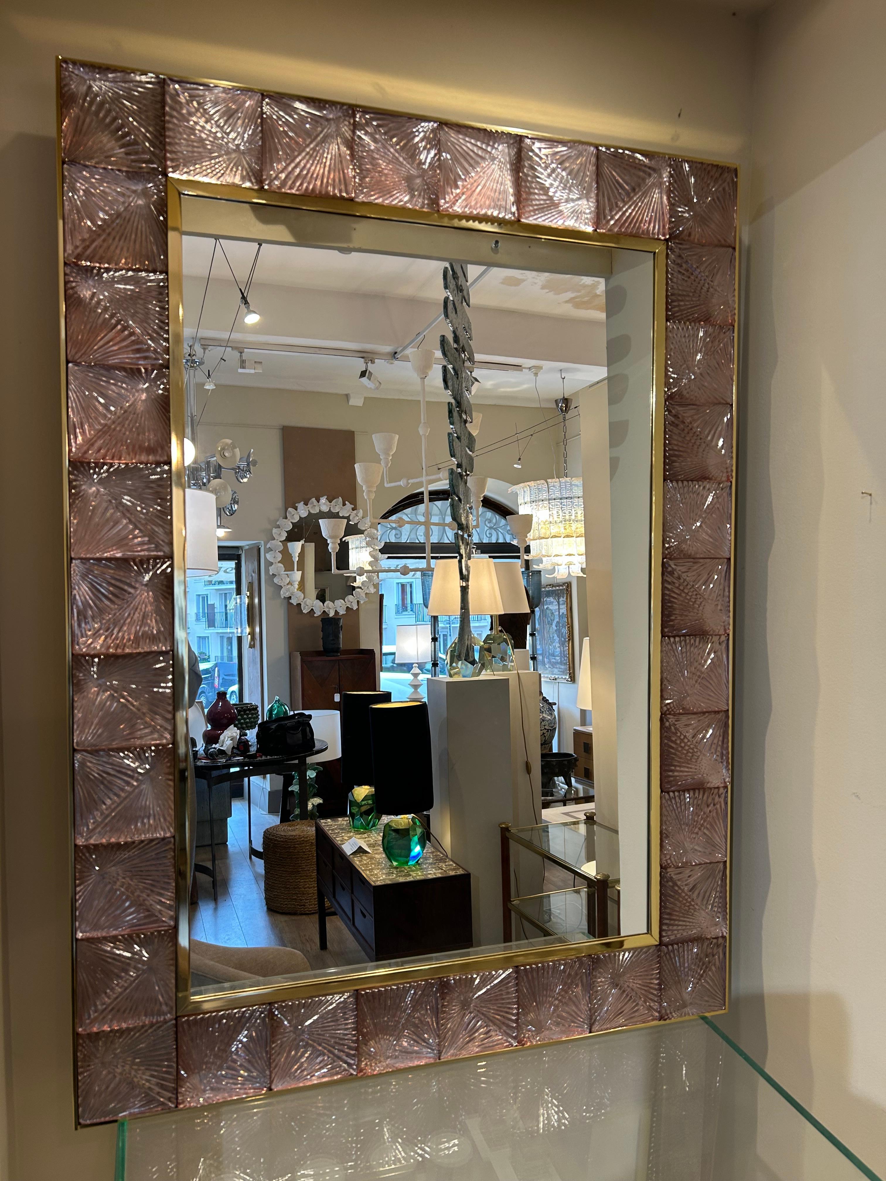 The mirror is rectangular in shape, framed by a decorative border made of textured Murano glass tiles. Each tile features a raised pattern forming a sort of fan or radiating starburst. The glass is pale pink in color and is set within a gilded brass