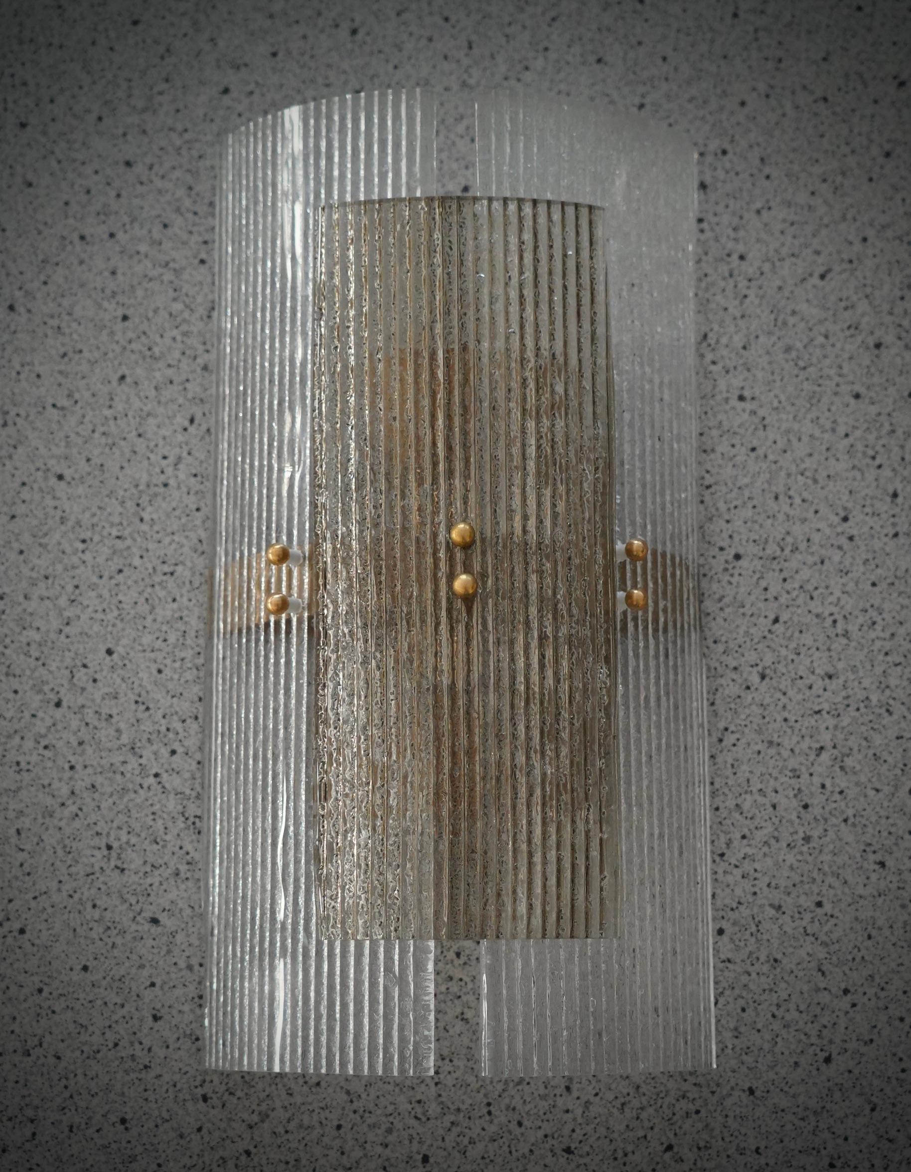 Este aplique de cristal de Murano presenta un bello montaje. Un cristal único y raro, con efecto ahumado y cristal transparente; estilo clásico de Murano.

El aplique está formado por dos hermosas piezas de cristal transparente estriado, curvadas