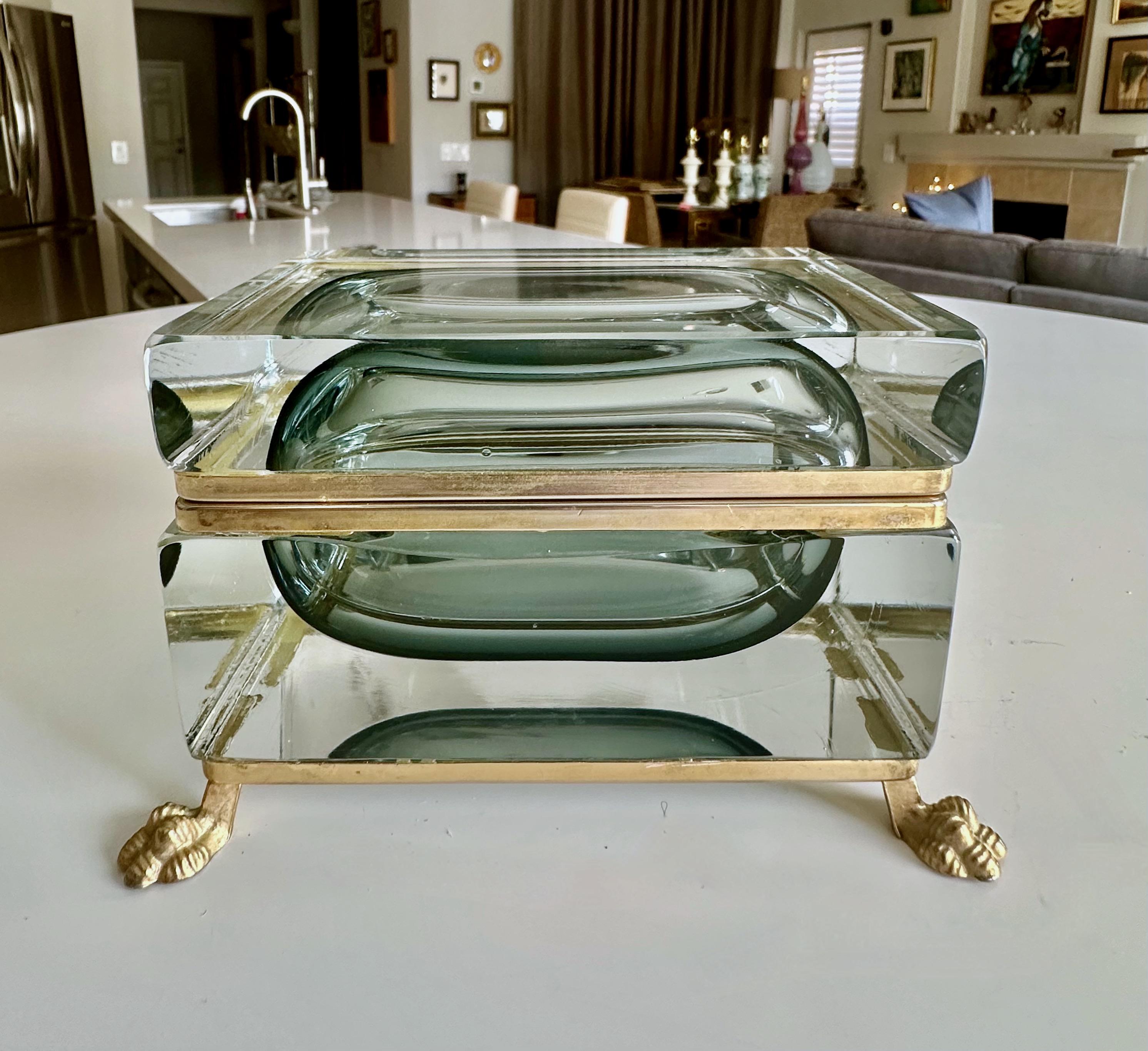 Caja cofre con tapa de cristal de Murano con marco de oro y latón que descansa sobre patas. La caja es octogonal de cristal transparente con hueco interior oval gris/verde.  Estampado en el marco de metal G.S.E. Ottone Garantito.