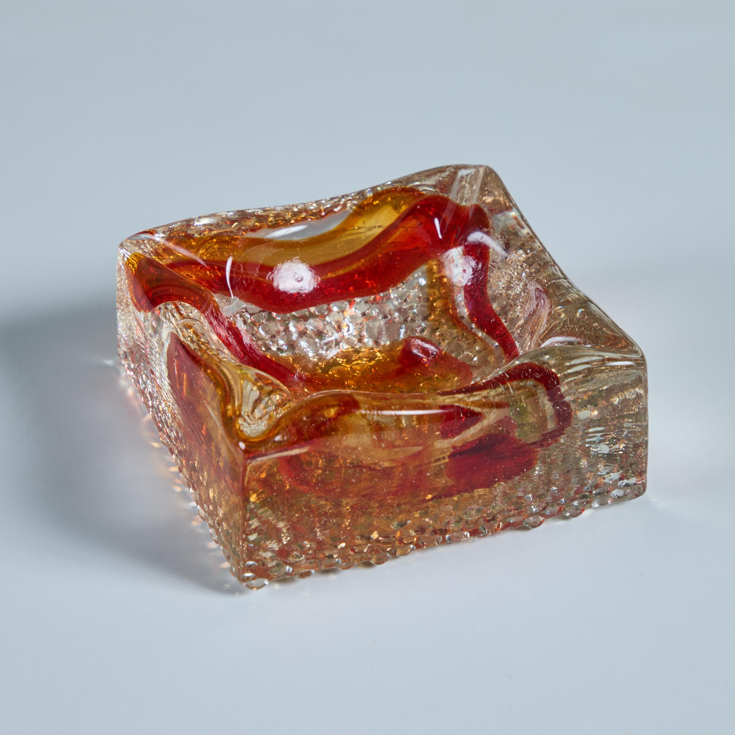 Ce cendrier du milieu du siècle dernier présente un corps carré en verre transparent épais avec un tourbillon orange rougeâtre sur toute sa surface, un puits central profond avec quatre fentes à chaque coin et un dessous texturé. Peut également être