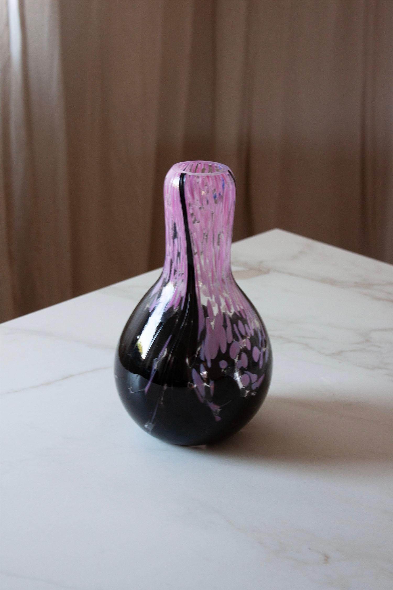 This hand-blown art glass vase features a beautiful interplay of deep black and soft pink hues, seamlessly fused in a fluid, organic form. The elongated neck gracefully tapers into a rounded body, while the pink detailing creates movement and depth