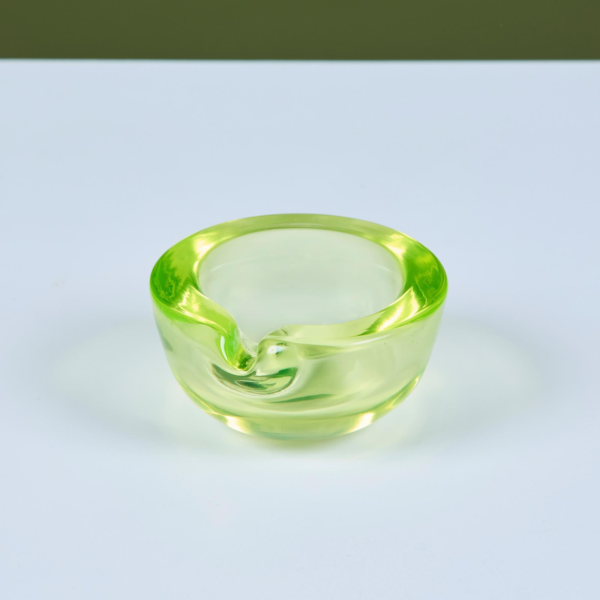 Runde Schale oder Aschenbecher mit Zwickel aus Glas. Das exquisite Uranglas, das in einem neonfarbenen Gelbgrün erstrahlt, setzt ein gewagtes Statement. Dieses skulpturale Stück aus dickem Glas kann als Videdose, Dekoschale, Briefbeschwerer oder
