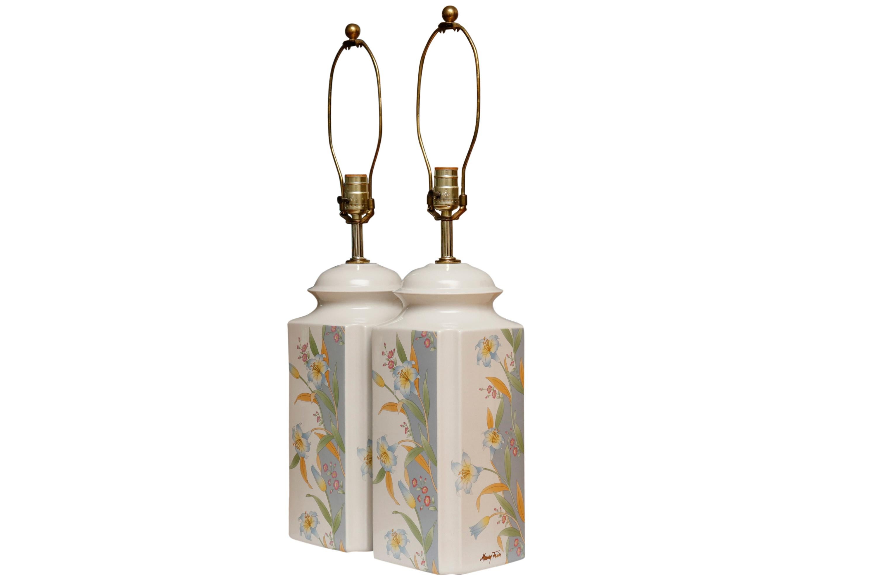 A pair of signed Murray Feiss table lamps. White ceramic vases are squared with concave curved corners. Printed in front with a floral motif over a blue wave. Harps are topped with ball finials made of brass. Wired and working. Each lamp measures