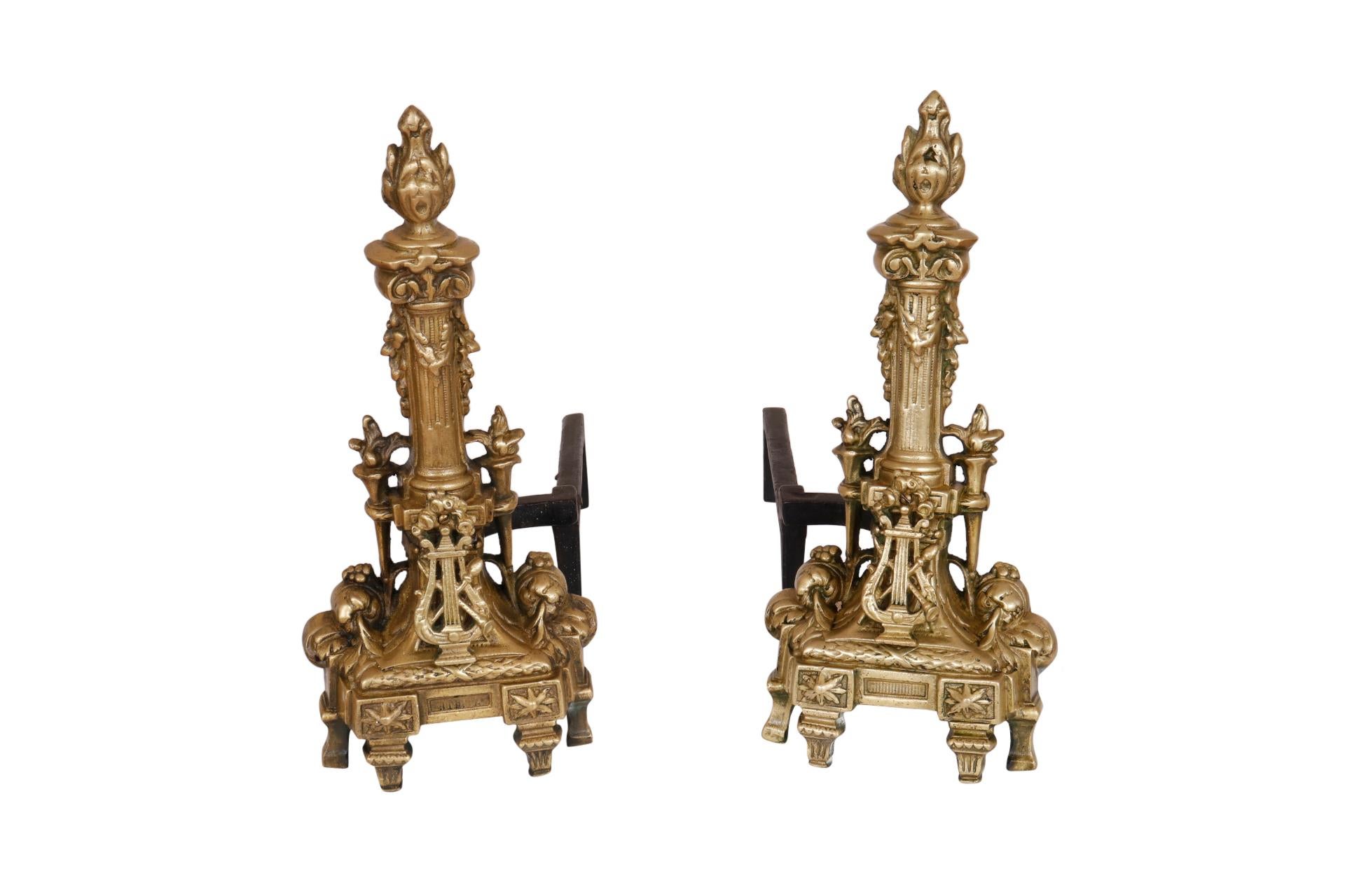 A pair of Regency style andirons by United Manufacturing. Brass and iron andirons are decorated with musical details. Central columns shaped like candles with acanthus swags are flanked with small torches. In front, a lyre is crossed with a clarinet
