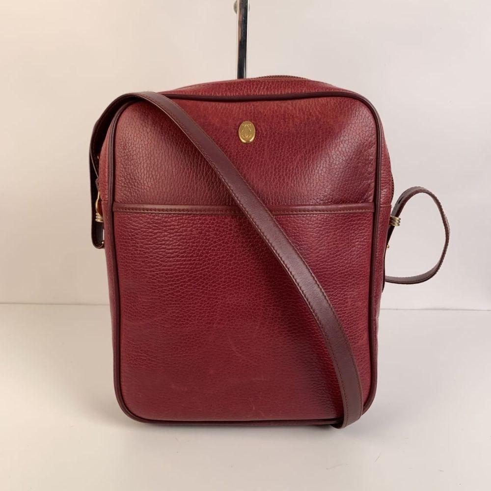 Vintage Must de Cartier Messenger bag crafted in burgundy leather. Upper zipper closure. Gold metal CC logo on the front. 1 open pocket on the front and 1 zip pocket on the back. Adjustable shoulder strap. Burgundy fabric lining with CARTIER logo's