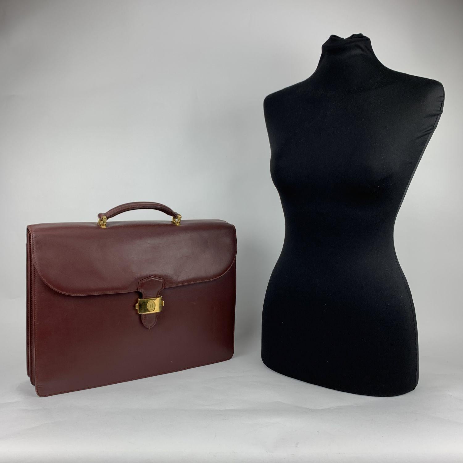 Classic and elegant Briefcase work bag by Must de Cartier. Crafted in smooth burgundy leather. Gold metal hardware. Flap closure with press lock closure on the front. Rear open pocket. 2 main internal compartments, with 1 side open pocket and 3 pen