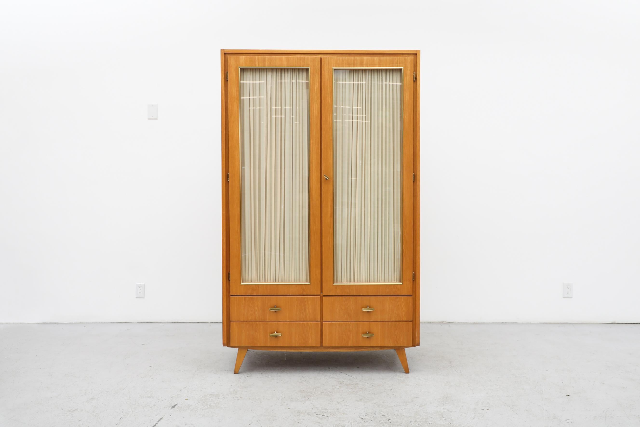 1950's Musterring locking wardrobe with locking glass doors and 4 lower drawers. The wardrobe has cream curtains on the inside of the glass doors and a brass toned clothing rod. Blonde exterior with mahogany interior. This piece is in good original