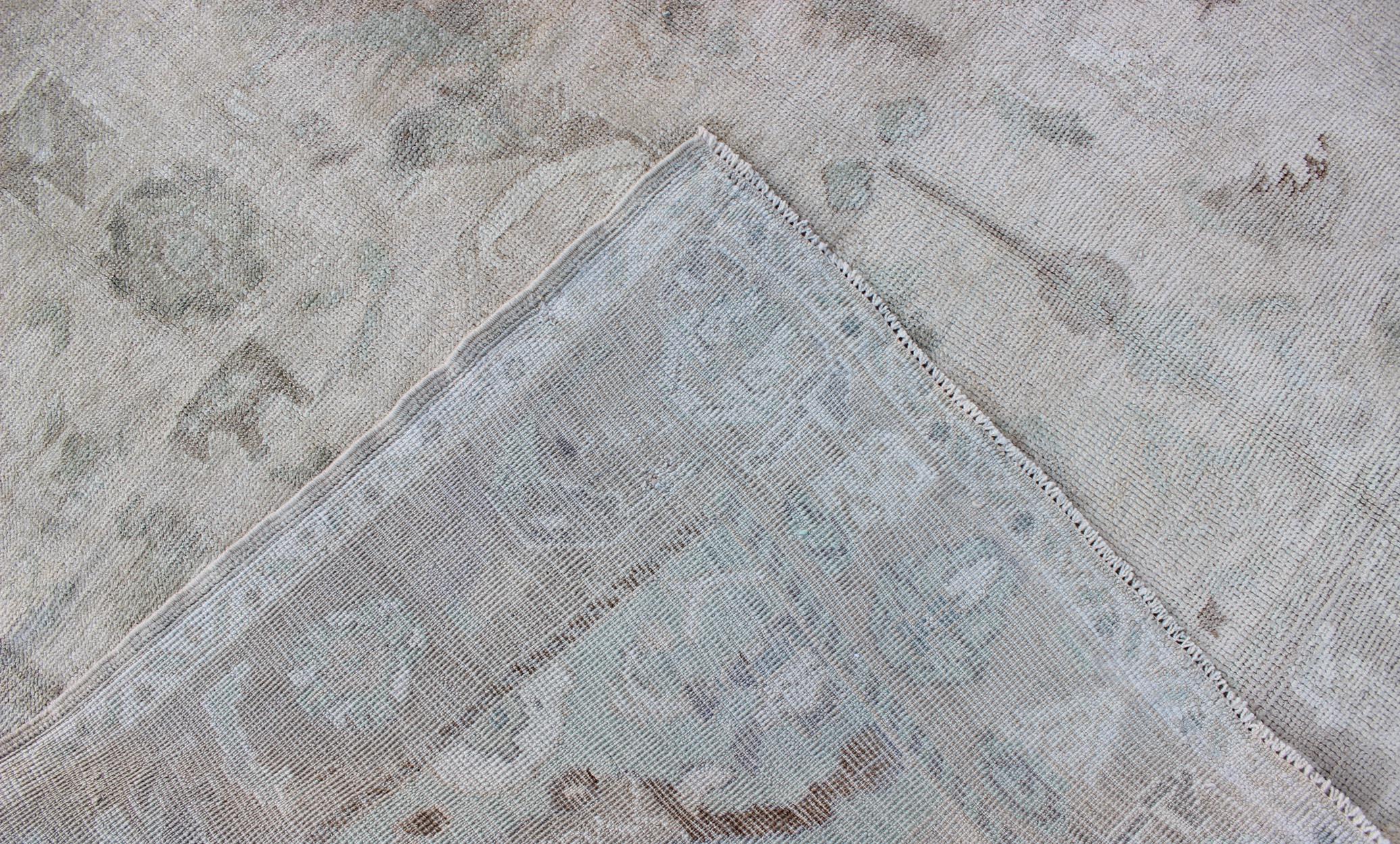 This Oushak rug features a unique blend of colors and an intricately beautiful design. The multi-layered Sub-geometric floral medallion is complemented by four similarly designed cornices. The various shades of cream, light blue, taupe, and brown