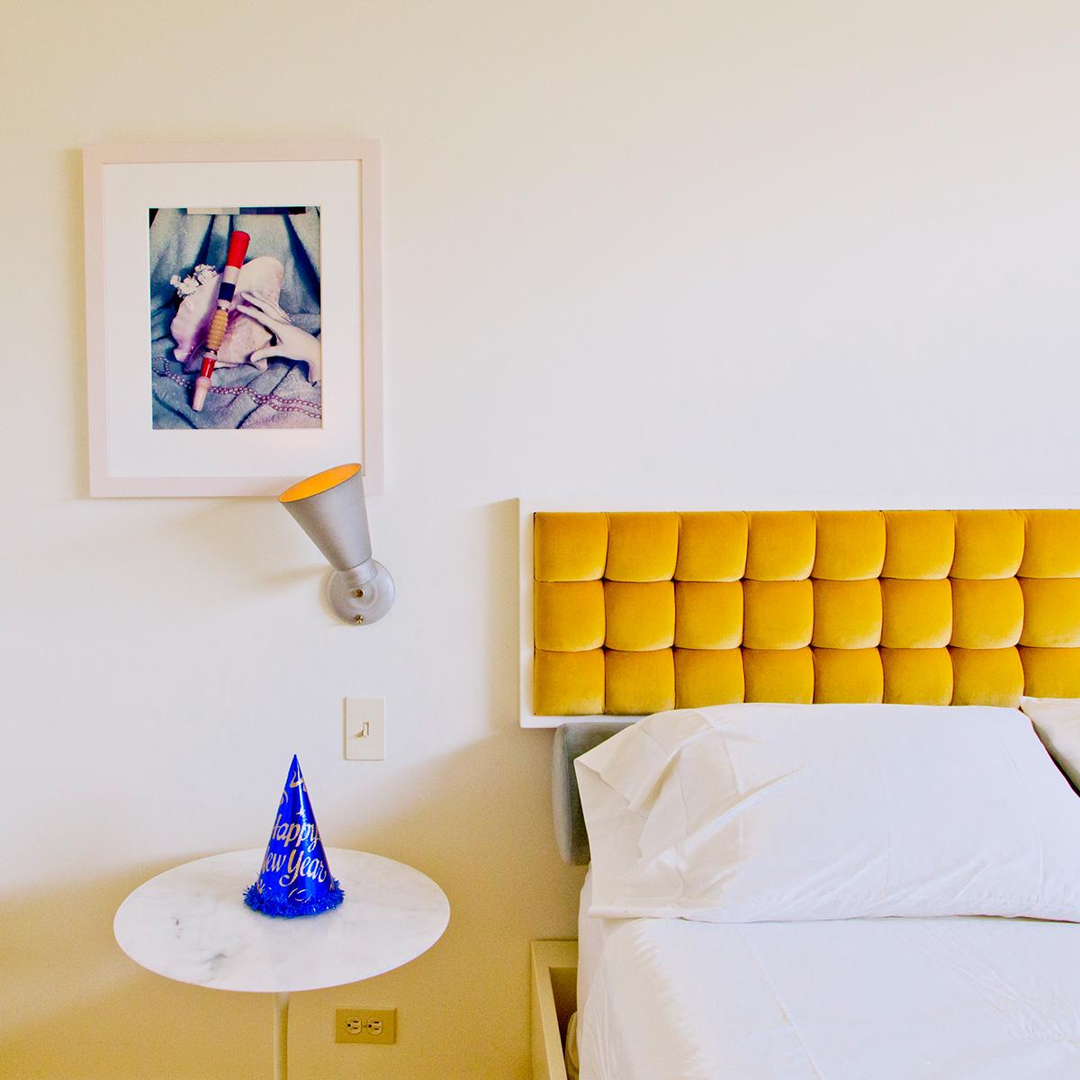 Happy New Year by Nancy Baron is a 16.5 x 16.5 inch archival pigment print. This photograph is available in an edition of 10. The photograph features the interior of a mid century modern bedroom, with a yellow headboard and a "Happy New Years" party