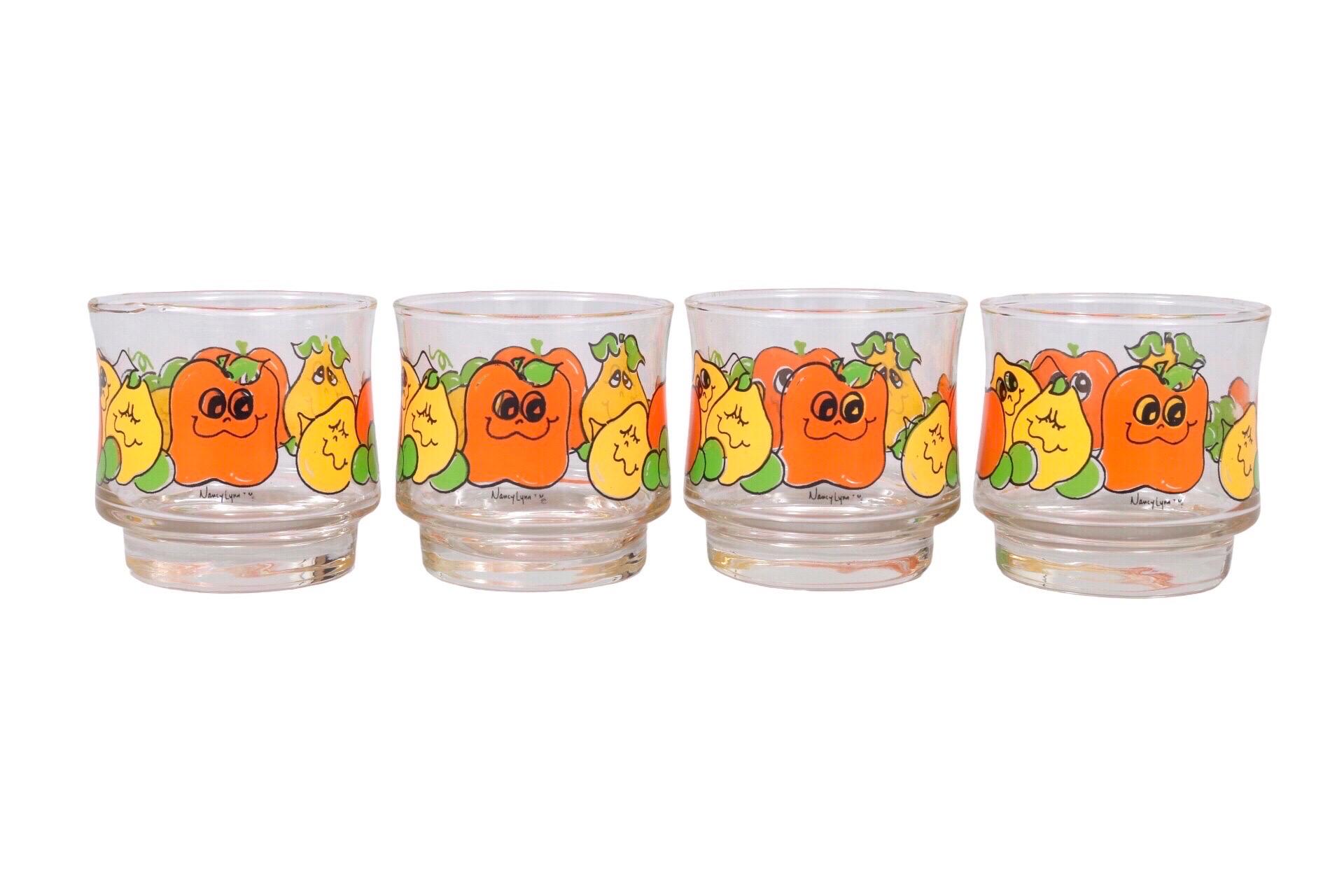 A set of four Nancy Lynn for Anchor Hocking glasses. Concave curved glasses for easy holding are decorated with stylized green, yellow & orange fruits. Lemons and apples have smiling cartoon faces. The design includes the Nancy Lynn signature