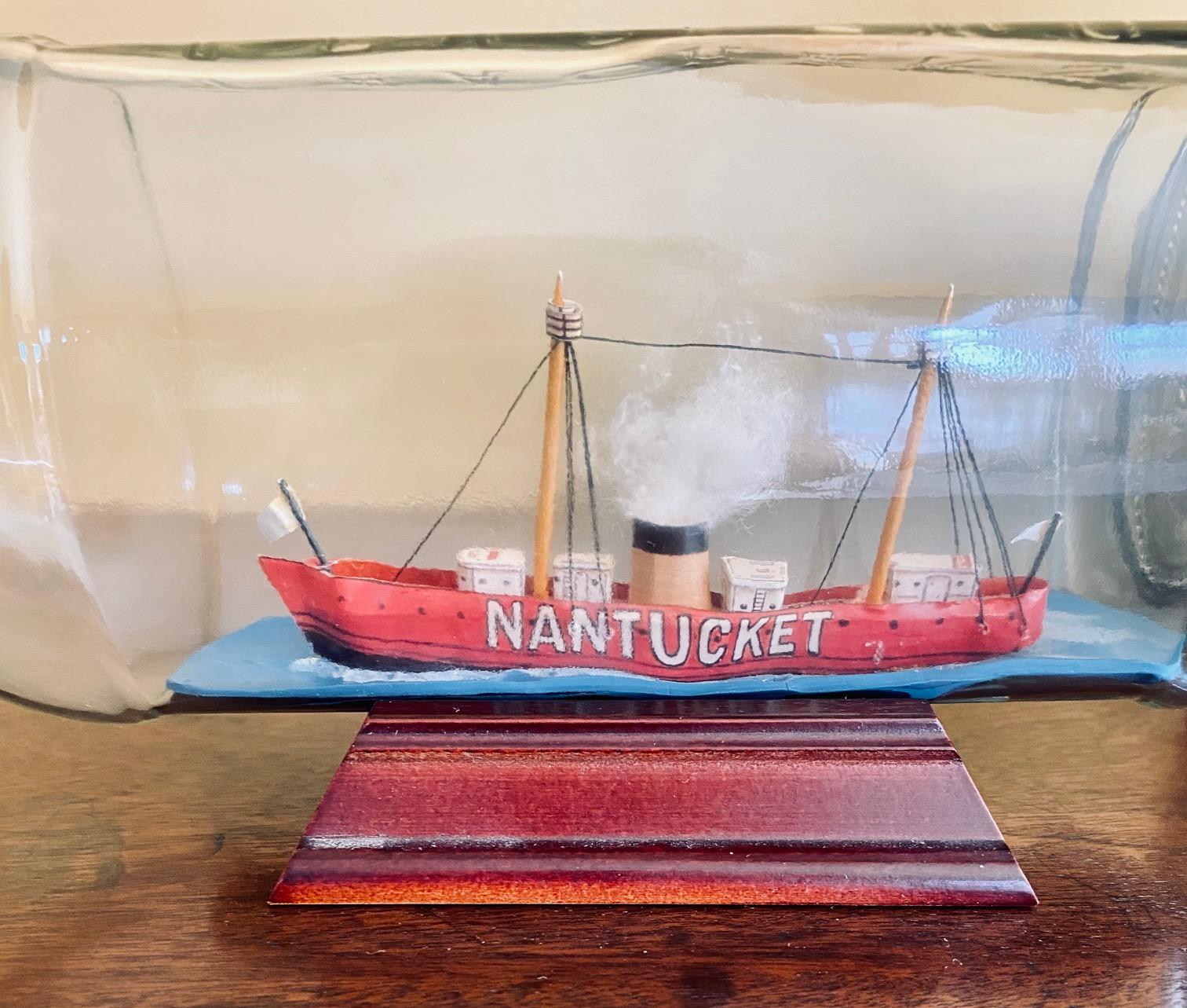 A delightful vintage ship-in-a-bottle depicting the Nantucket lightship, circa 1960, with a paper on carved wood hull painted in the Classic red with station name NANTUCKET on both sides in bold white letters; two masts supporting light beacons with