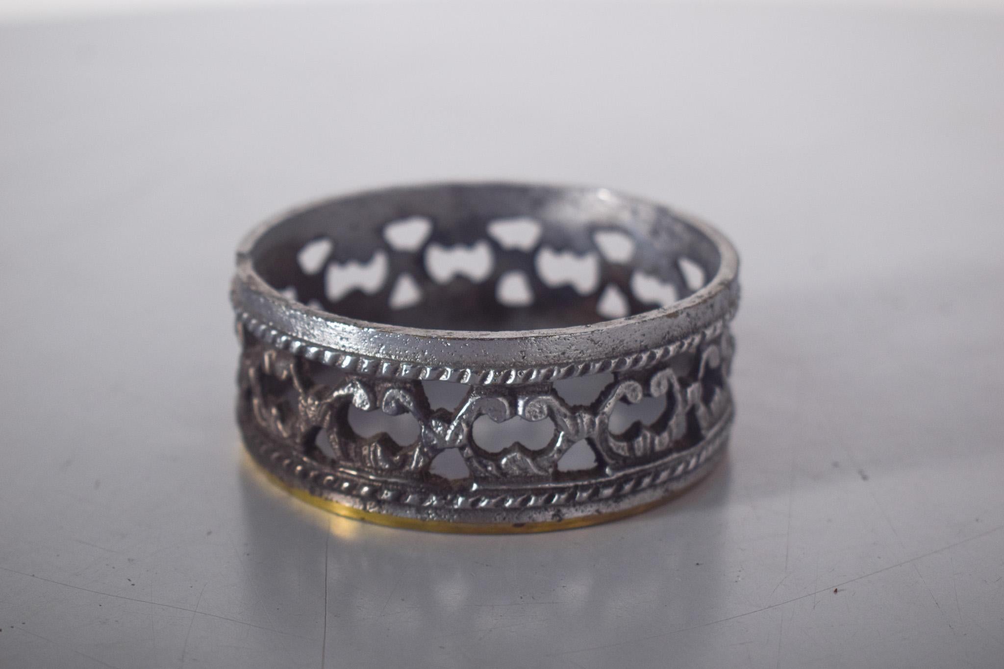 Rond de serviette en métal travaillé. De forme cylindrique et ajourée, avec de délicates découpes en motifs symétriques rappelant des arabesques, il présente un style sophistiqué et vintage. La finition vieillie met en valeur la texture de la pièce,