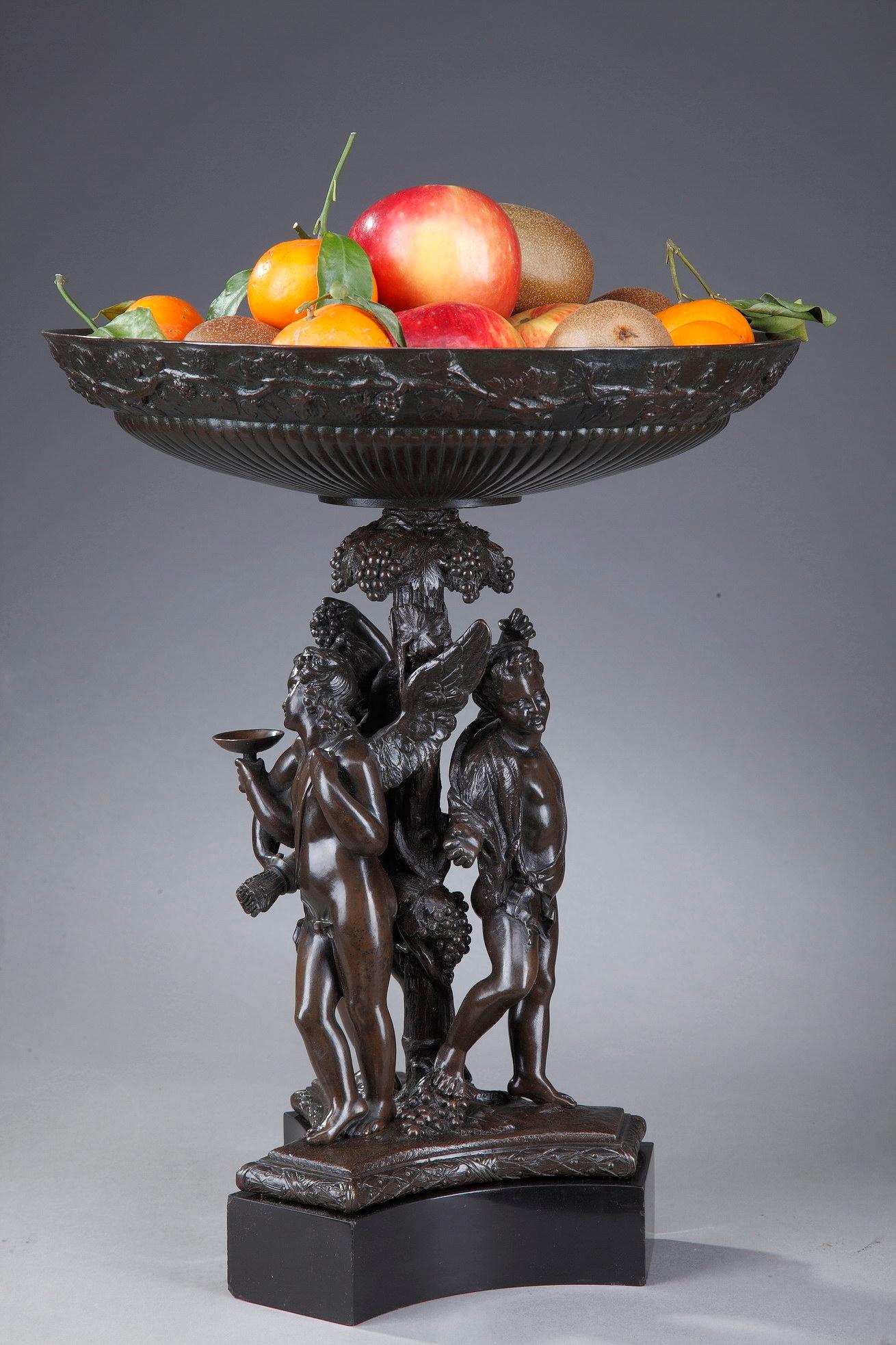 Carved of patinated bronze, this 19th century fruit bowl boasts a mythological scene with three winged Cupids wearing vine leaves, holding a cup and grapes. They are leaning against the central foot, which is richly decorated with vine leaves and