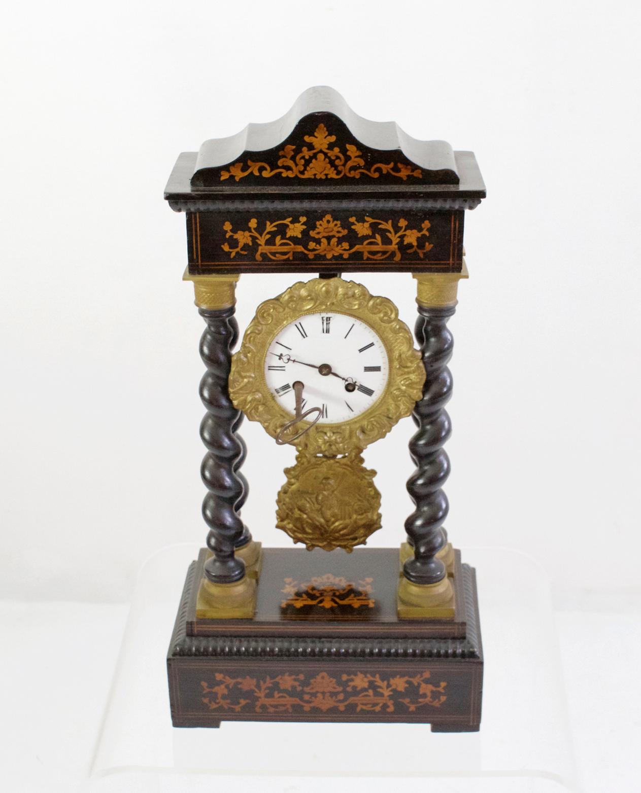 Mantle clock with an architectural design consisting of a stepped base with four columns and entablature with frieze and cornice. It is decorated with floral marquetry ornamentation and twisted columns attached in bronze casts. The dial has Roman