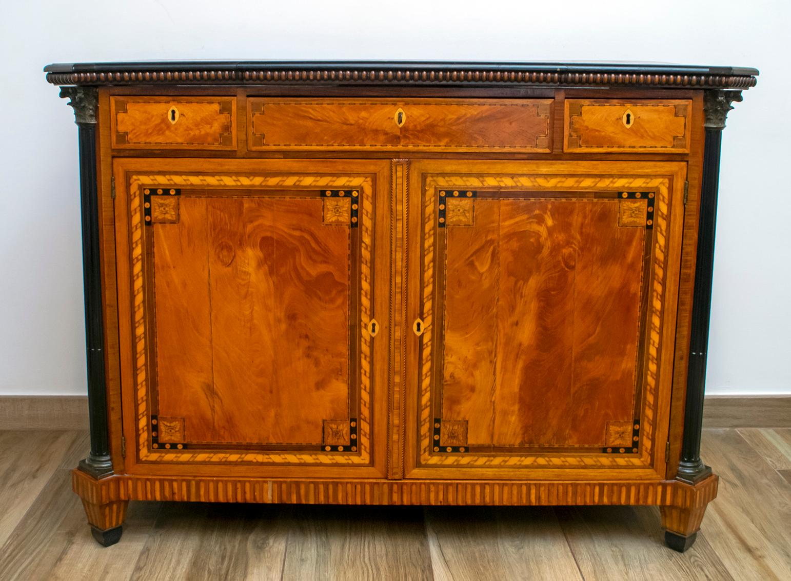 Elegant sideboard finely inlaid with floral and geometric motifs. It has three drawers and two doors with panels inlaid in various essences with geometric patterns. Enriched with capitals and column bases in gilded bronze. The opening top is inlaid