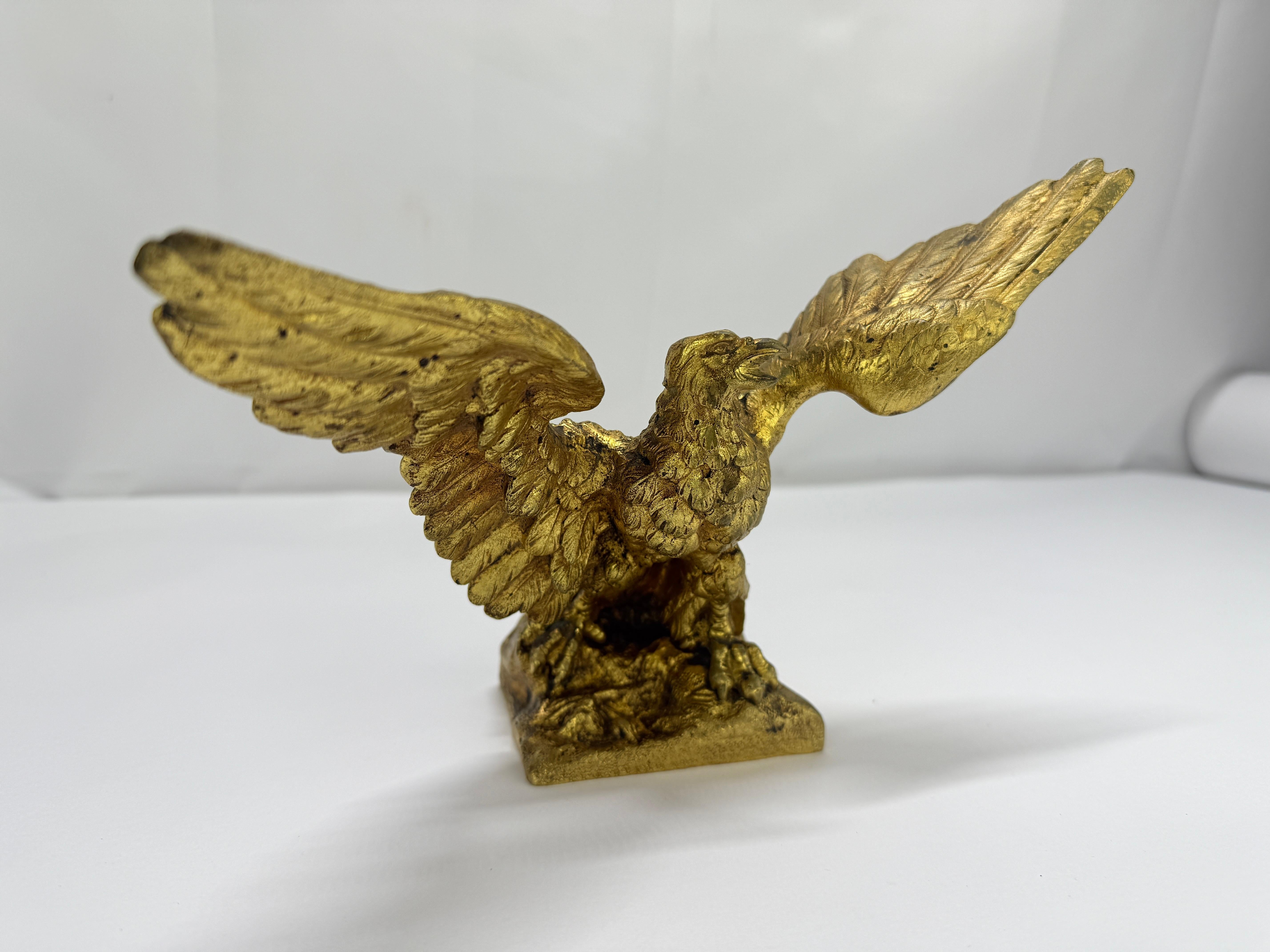 An animated, gilt bronze eagle arrests the moment of takeoff: shoulders thrust forward, primaries flared, and the head canted to the left with the beak parted in a cry. The composition is organized as a dramatic X—diagonal wings countered by the