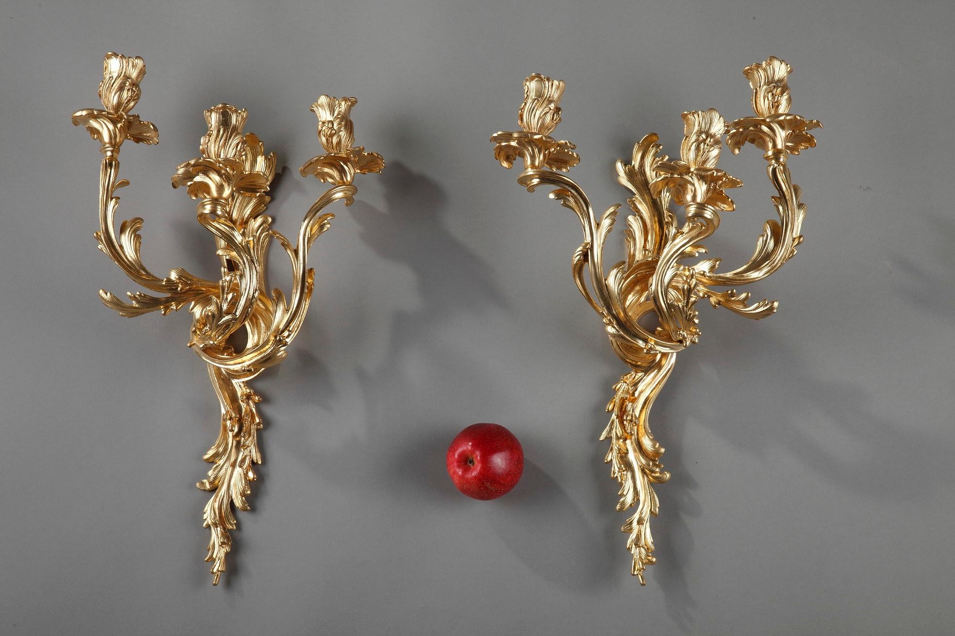 Pair of Rocaille sconces crafted in gilt and chiseled bronze with three-light. Richly decorated with flowing and asymmetrical foliage, these wall lights are a symphony of the Louis XV aesthetic, dominated by a plethora of realistic flowers, seeds