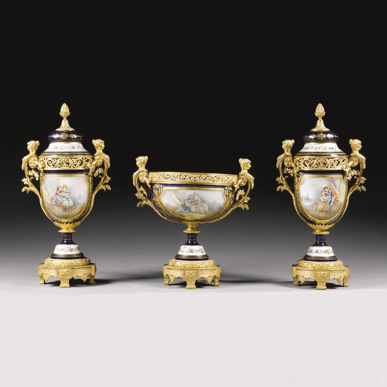 A Napoléon III gilt-bronze mounted Sèvres style garniture set. Comprising a centrepiece and a pair of vases with lids, by 'Robert'.

Each painted on the reverse with bouquets of flowers, to the front with romantic scenes, and mounted with bronze