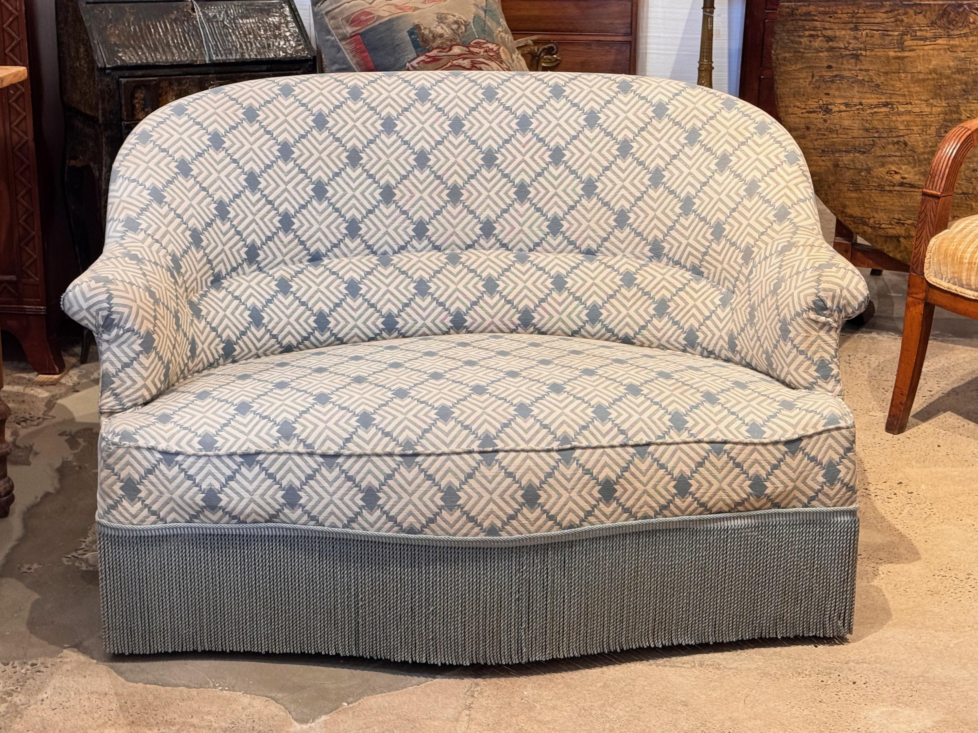 Compact upholstered settee in the Napoleon III style, featuring a rounded back, flared arms, and tailored silhouette. Upholstered in a geometric patterned fabric with a full fringe skirt. Works well as an accent piece in a bedroom or living area
